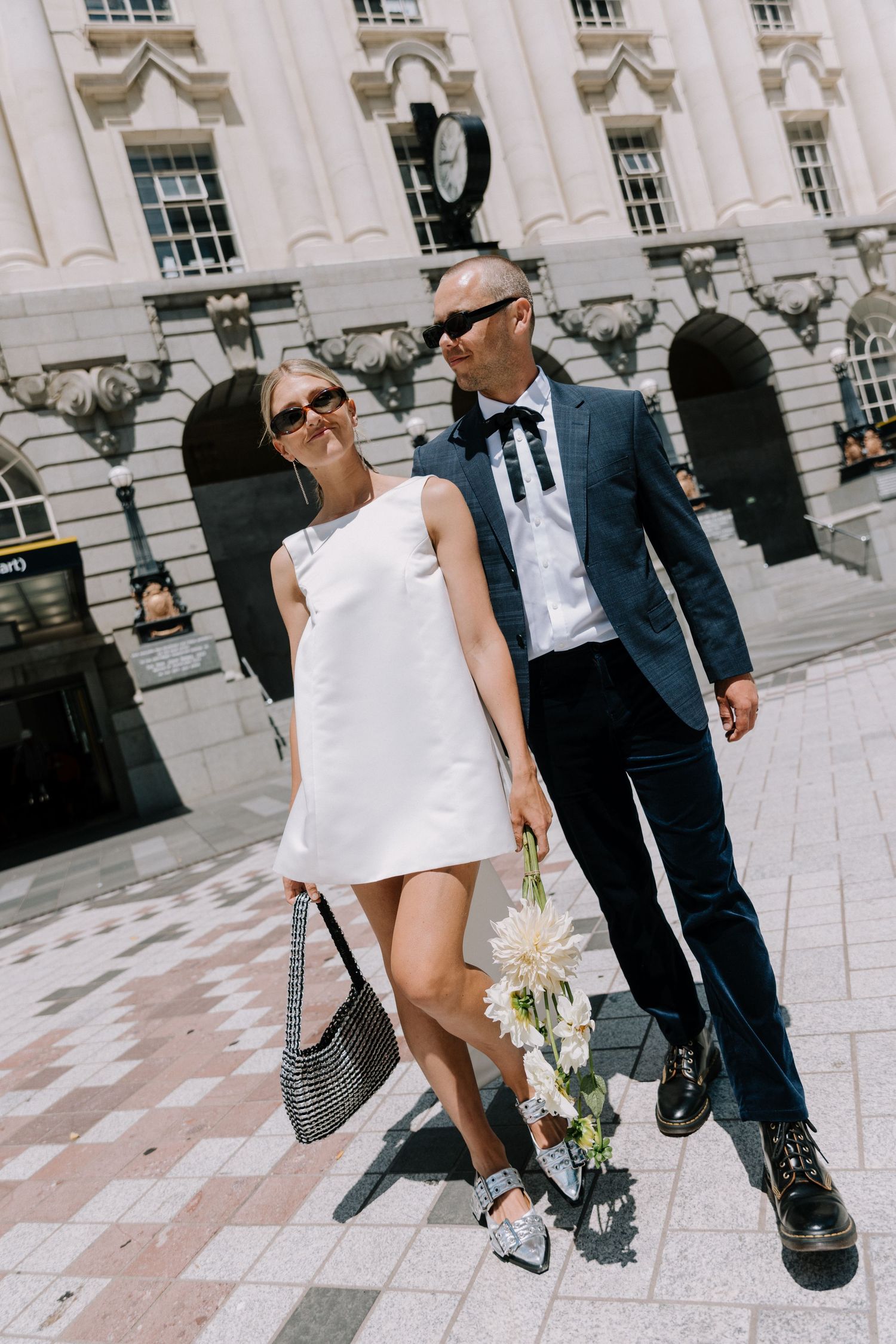 Stylish couple in wedding attire with sunglasses walking outside grand building on sunny city street.