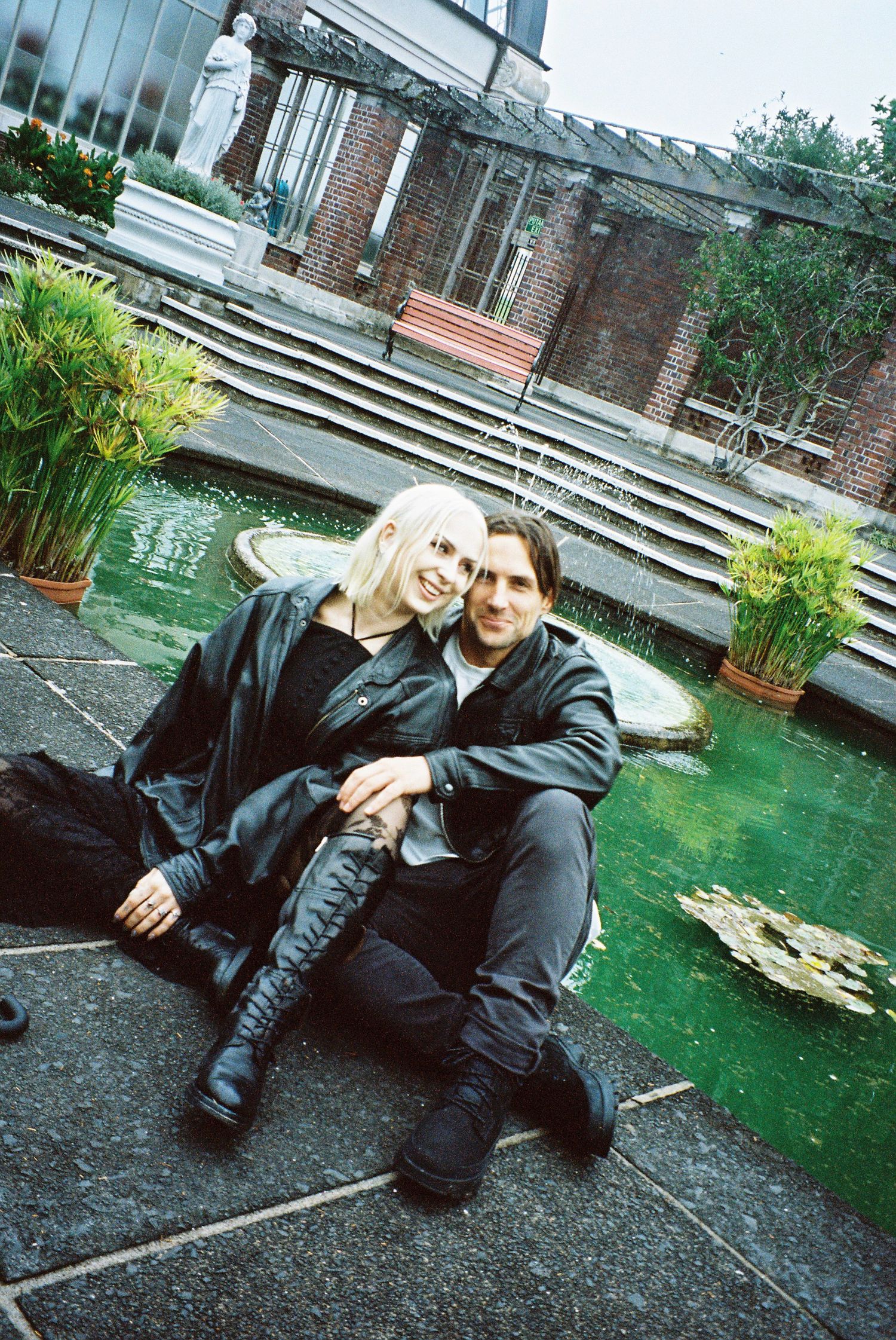 Couple in black outfits sitting together beside a green lily pond in a courtyard.