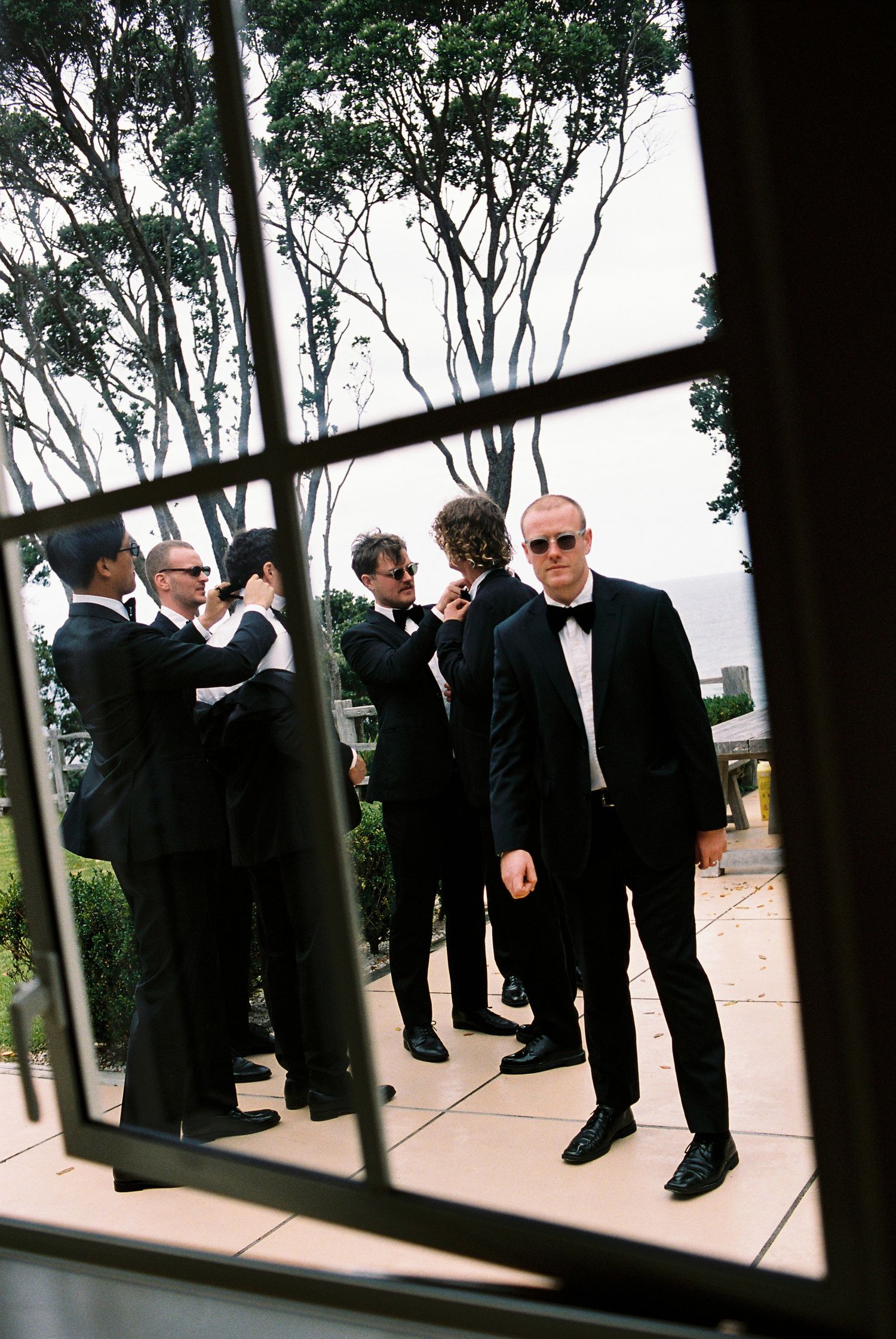 Groomsmen in black tuxedos prepare before wedding, photographed through a window reflection.