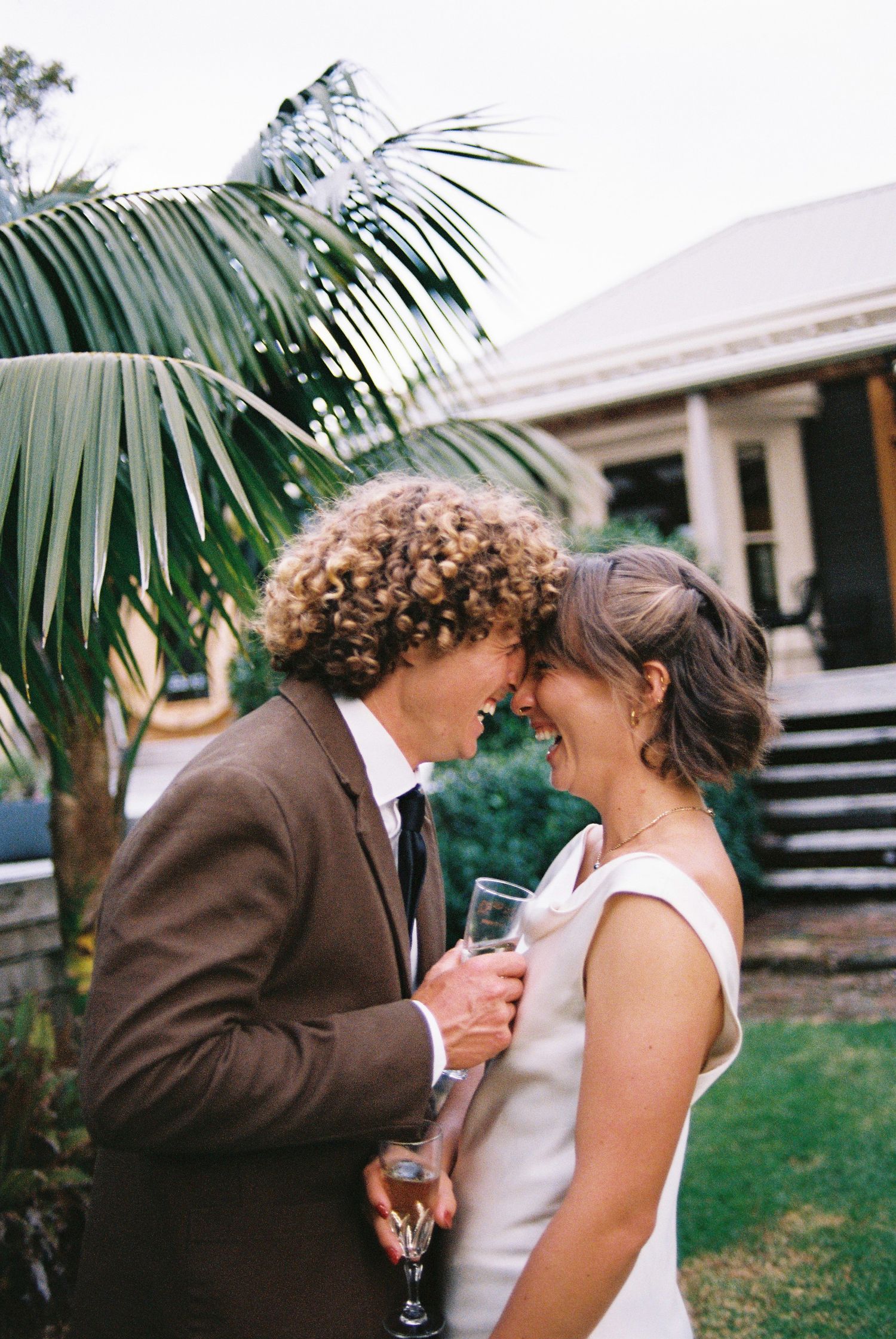 Joyful bride and groom touching foreheads and sharing champagne beside tropical palm tree.