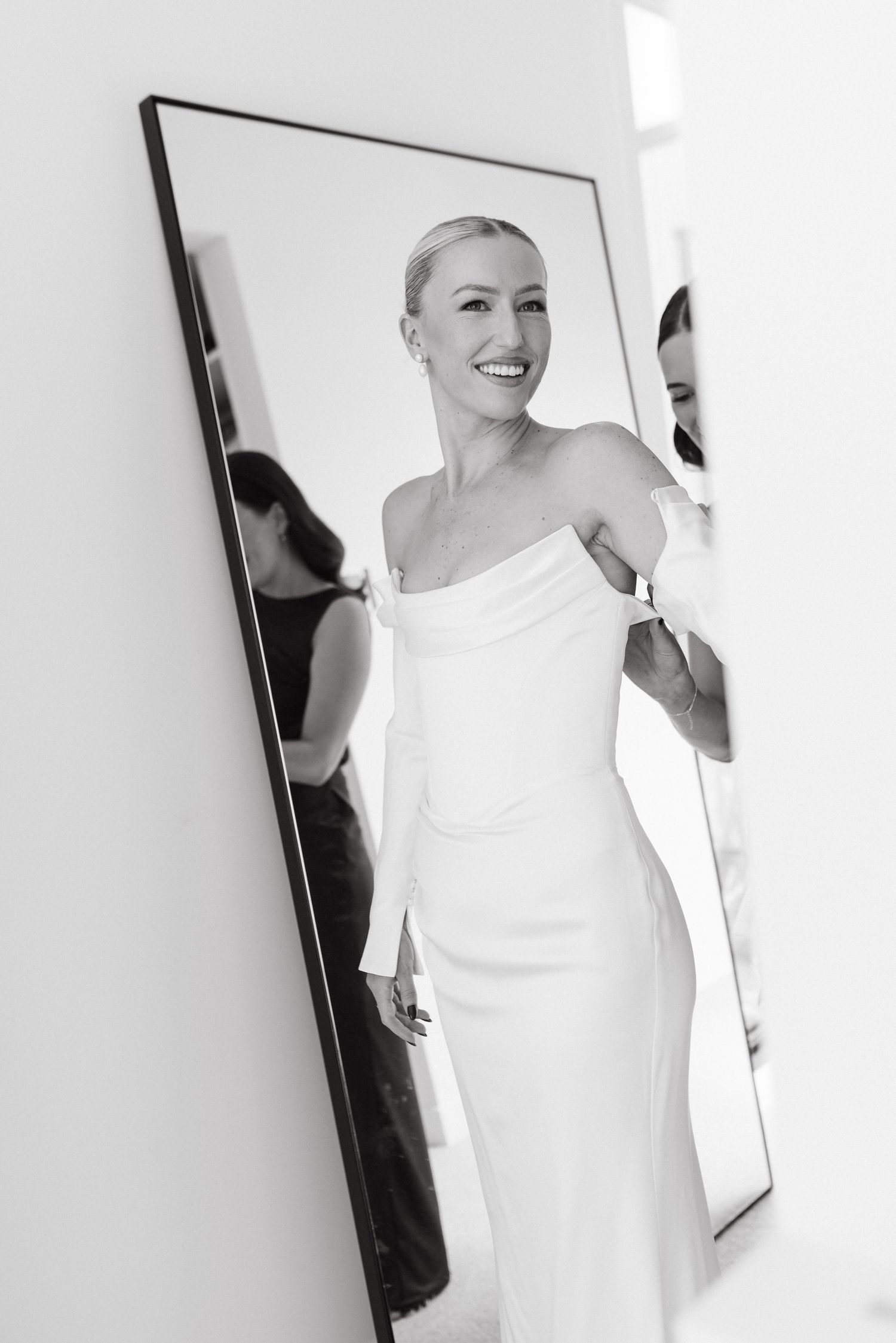 Black and white mirror reflection of smiling bride in off-shoulder white gown being dressed by assistant.
