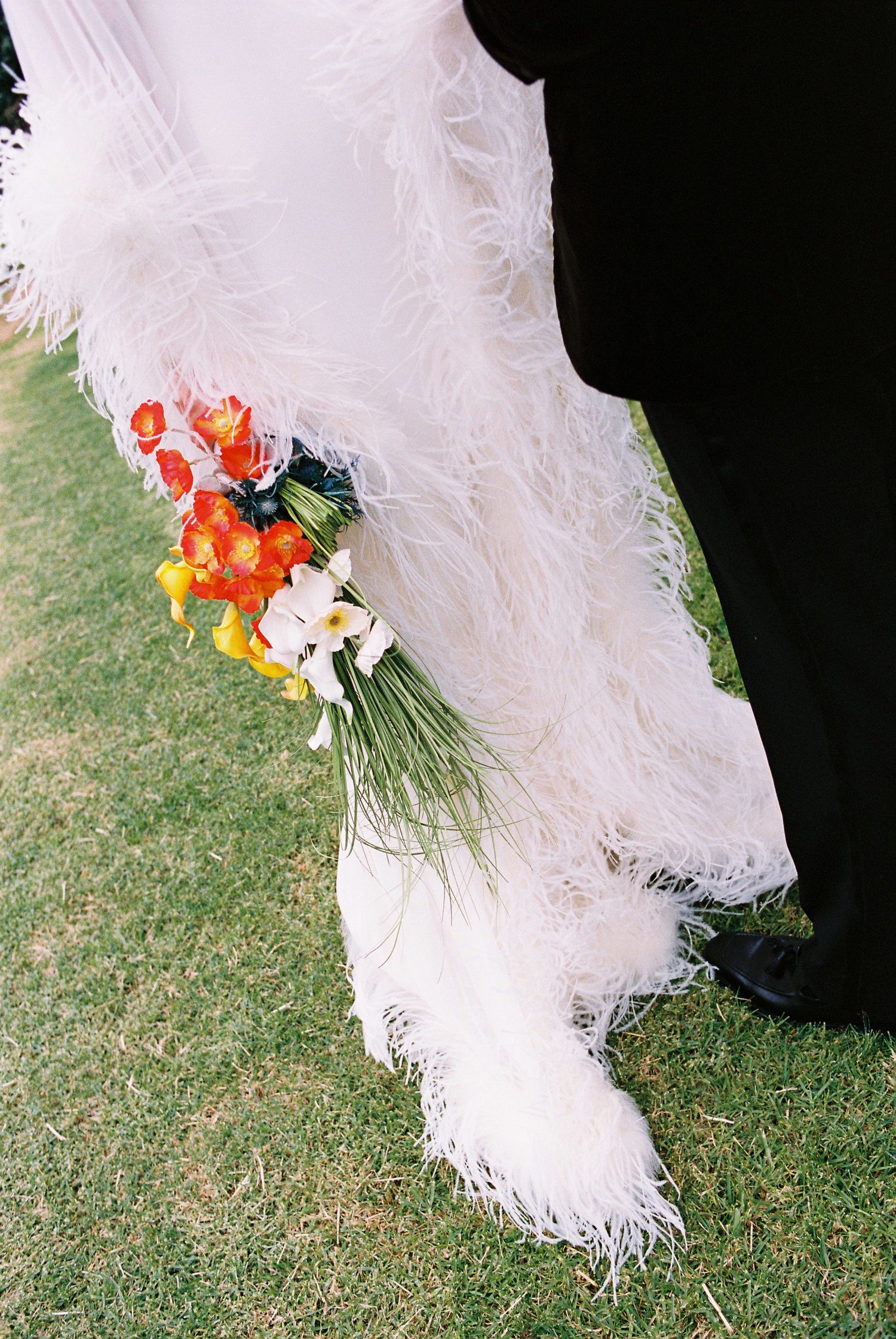 Close-up of bride's white feathered wedding dress hem and colorful bridal bouquet on green grass.