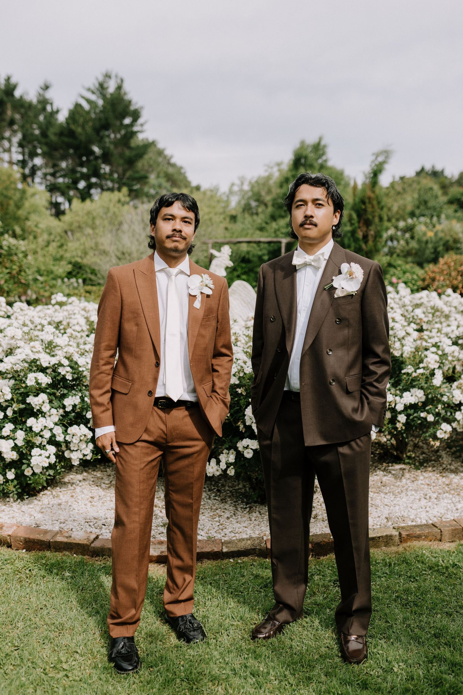 Two grooms in brown suits with boutonnieres pose in a garden with white flowers.