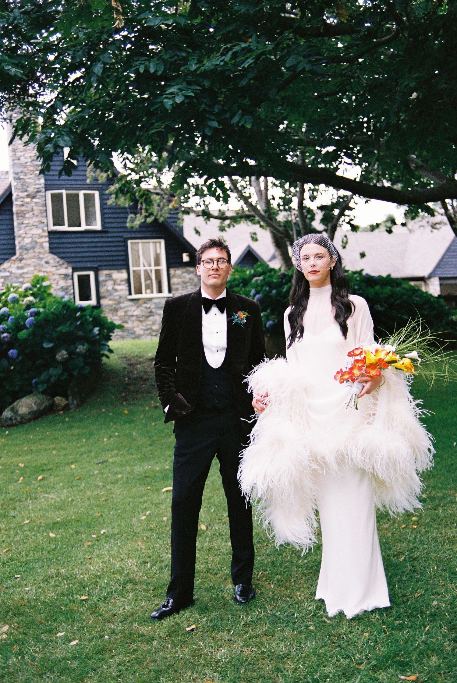 Bride in feathered white gown holds orange bouquet beside groom in black tuxedo outdoors.