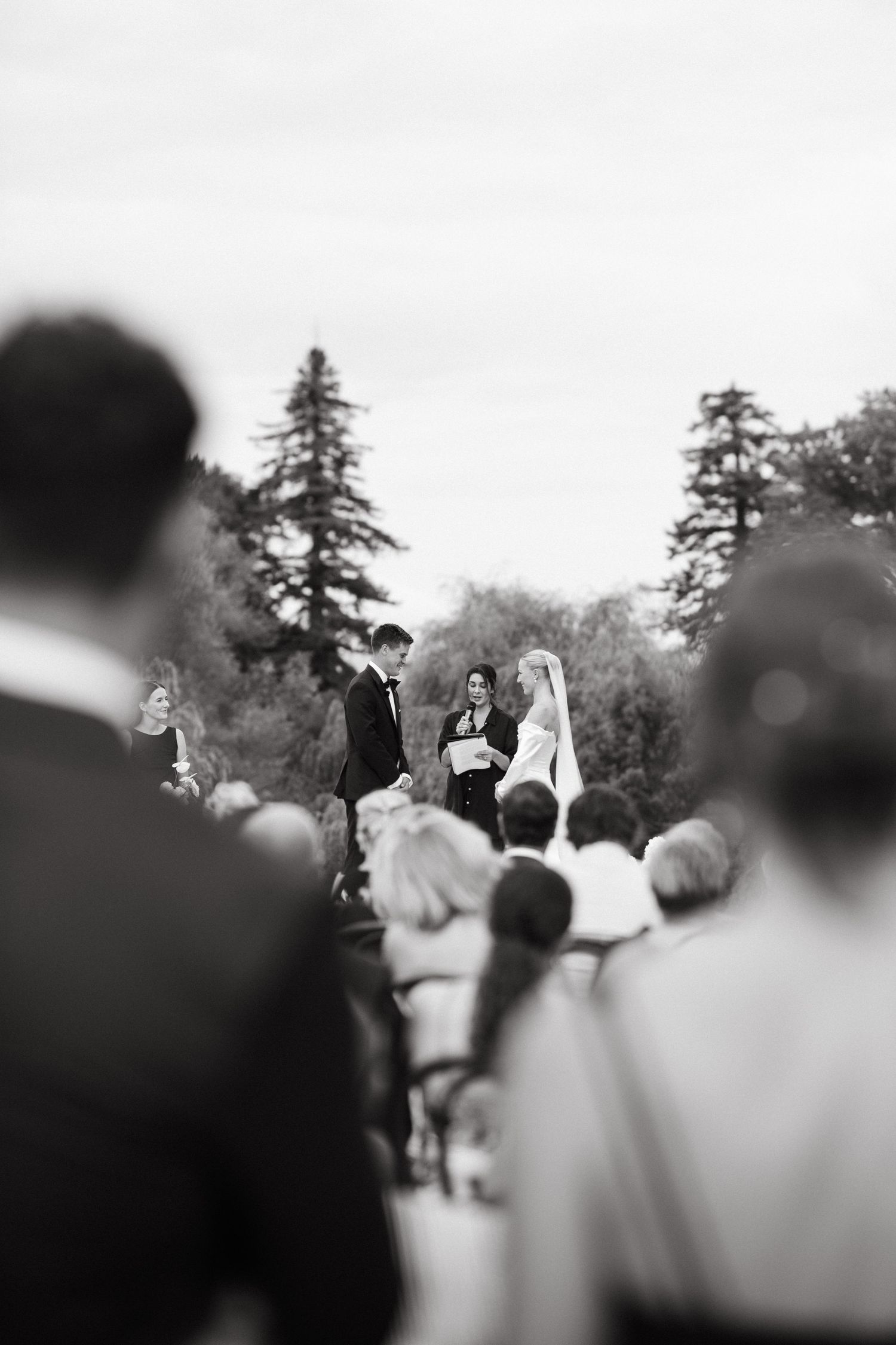 Black and white distant view of bride and groom exchanging vows at outdoor wedding ceremony with guests.