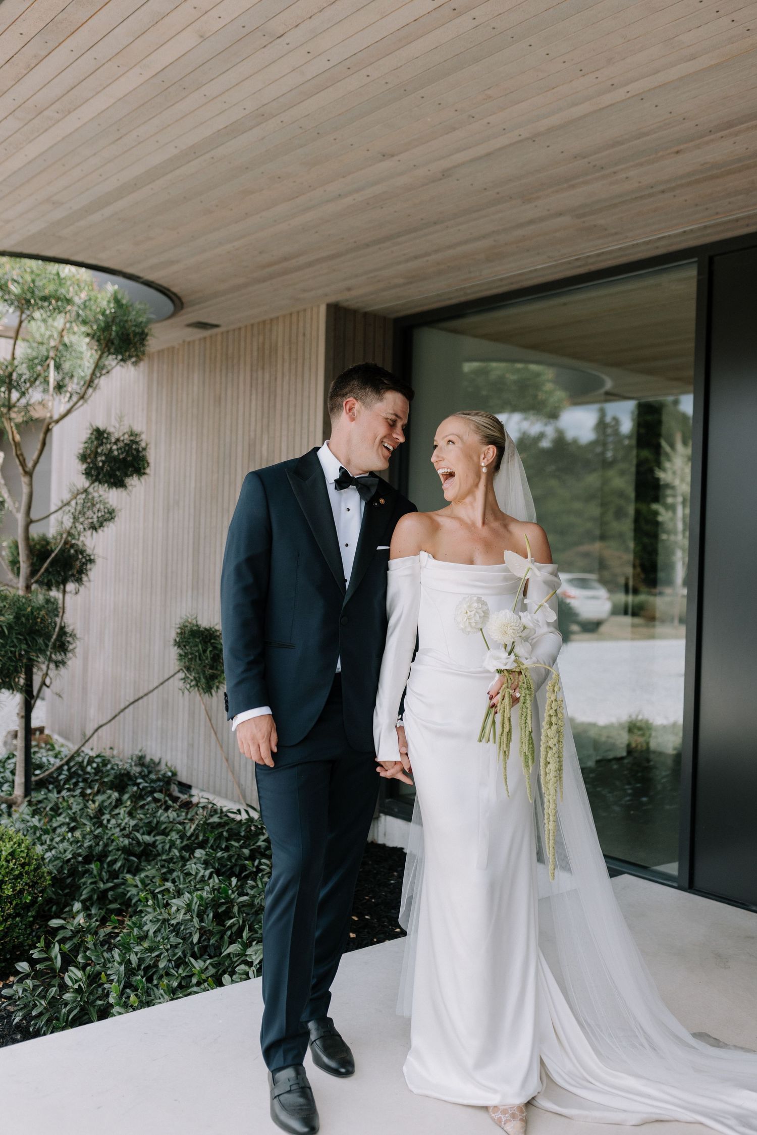 Bride and groom laughing together outside a modern venue, bride in off-shoulder white gown holding white bouquet.