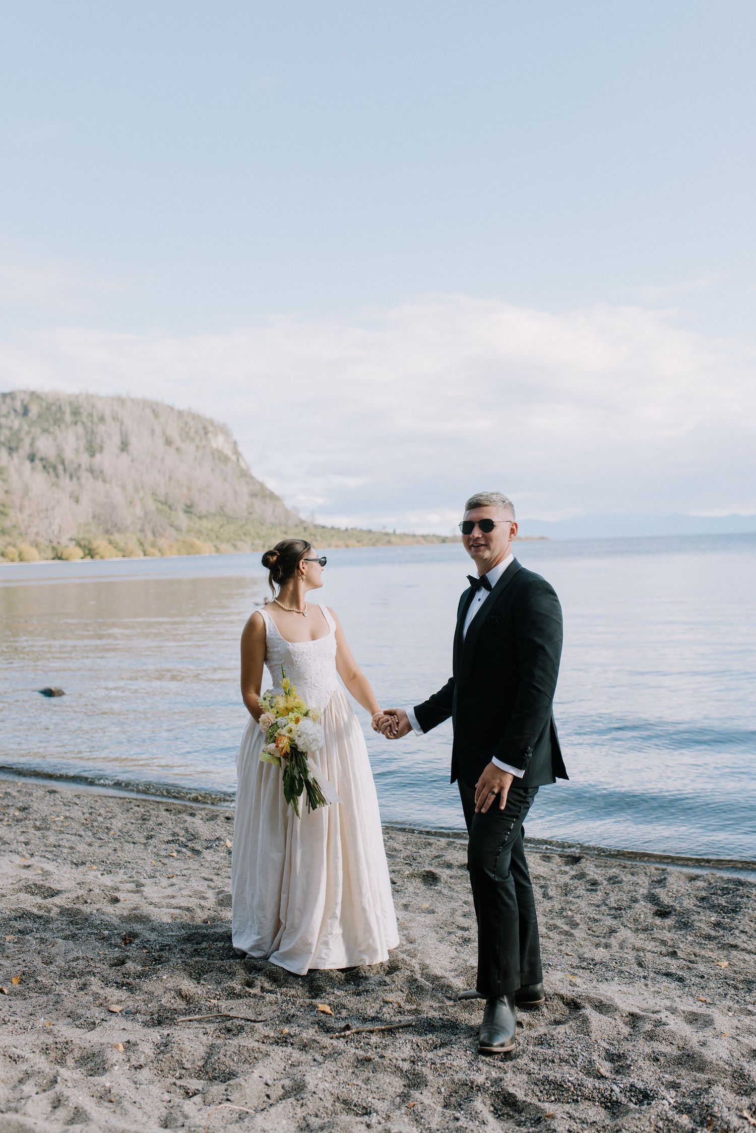 Bride and groom in sunglasses hold hands on a scenic lakeside beach with mountain backdrop.