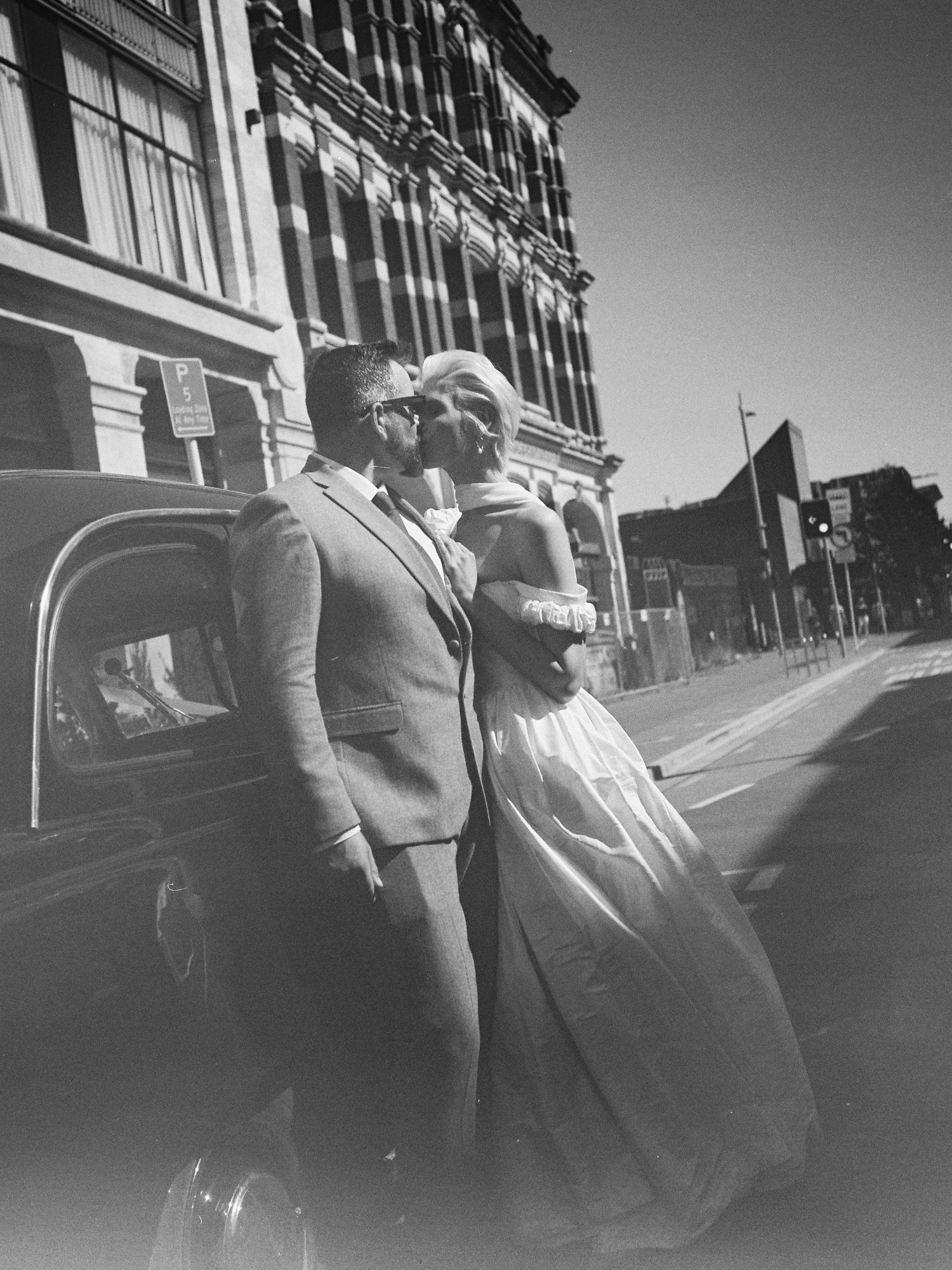 Black and white photo of bride and groom embracing beside a vintage car on a city street.