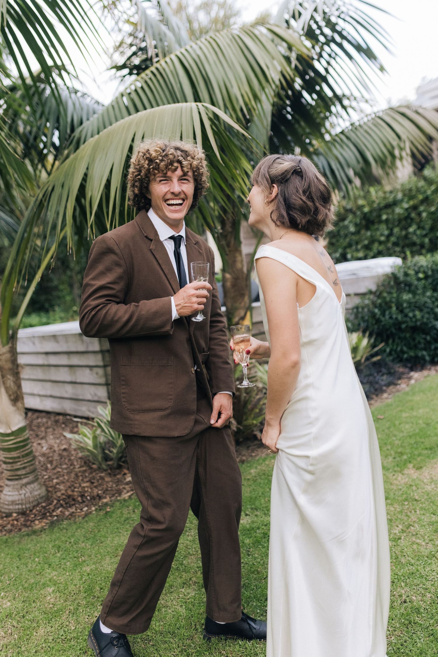 Laughing groom in brown suit holding champagne with bride in white dress in garden.