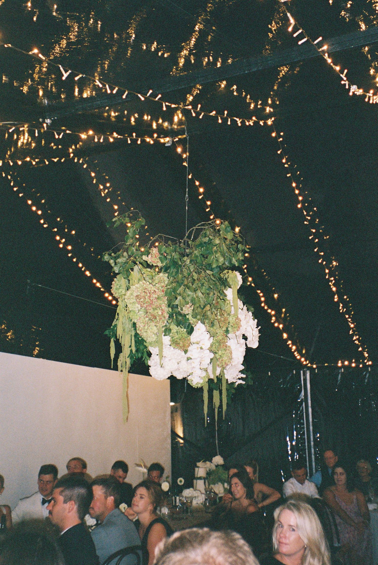 Hanging floral chandelier of white hydrangeas and greenery above wedding reception guests under fairy lights.