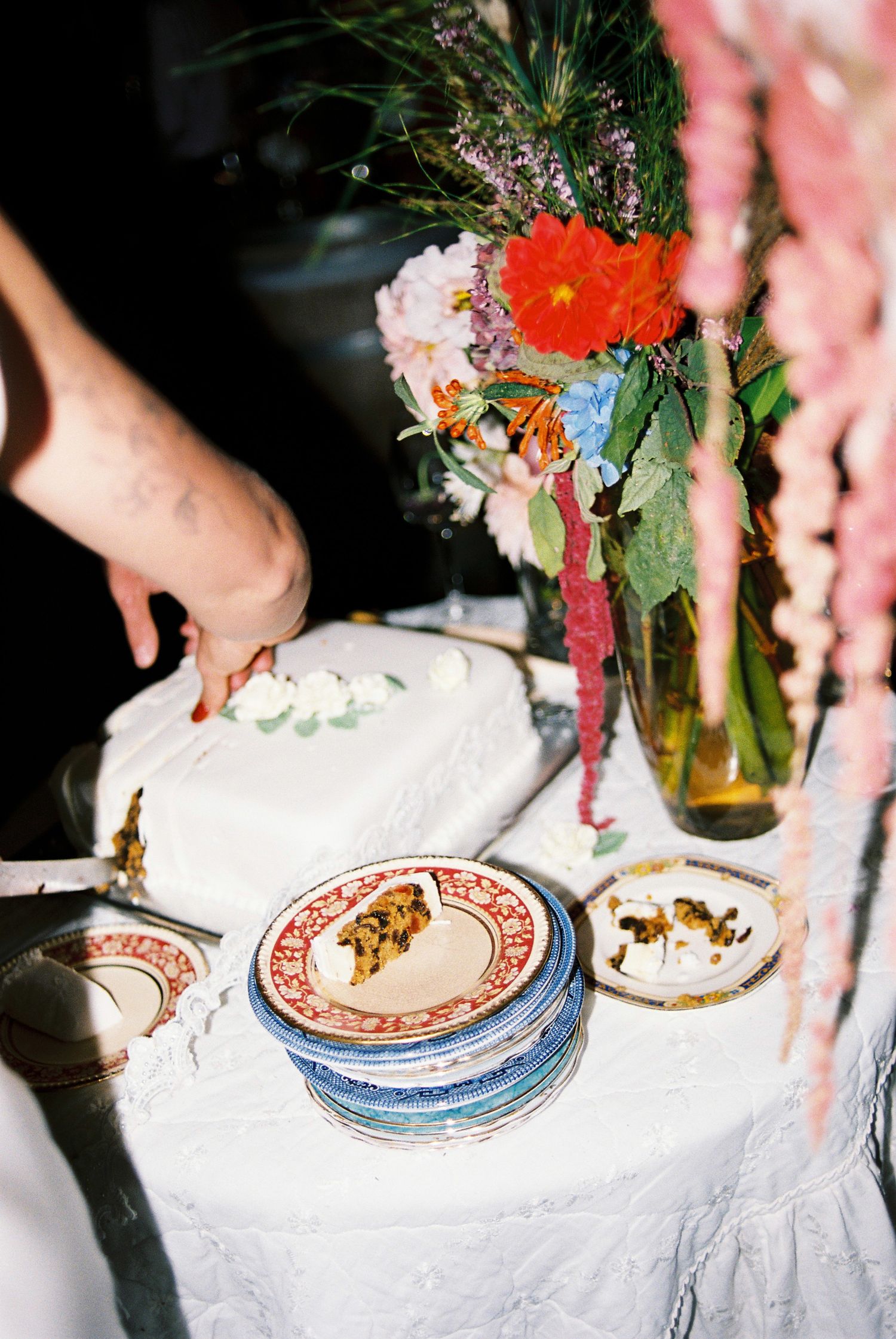 White wedding cake being cut on a table with colorful floral arrangements and vintage mismatched plates.