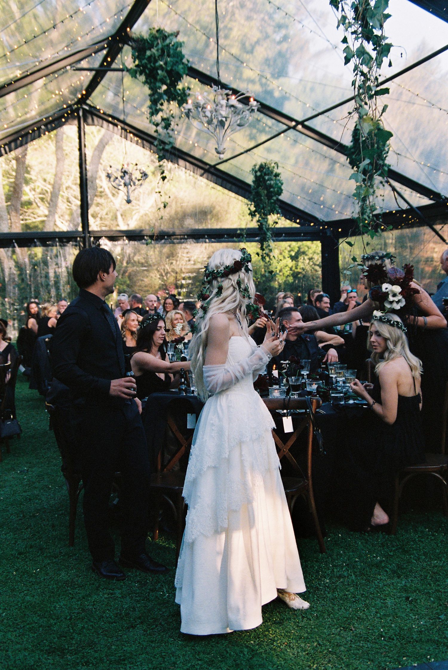 Bride in white tiered gown with floral crown celebrates with guests under a clear tent reception.
