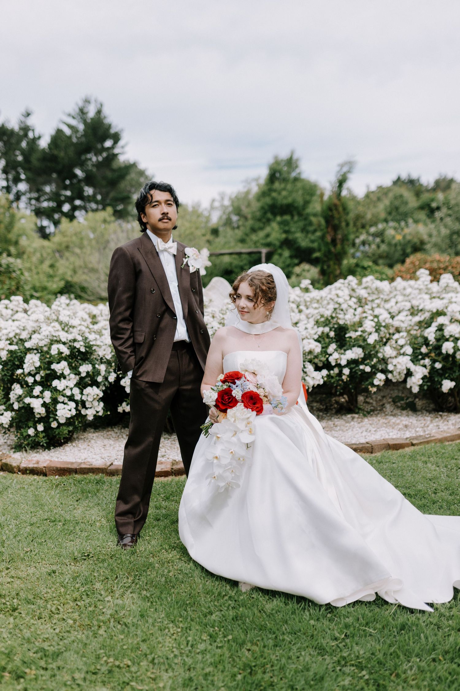 Bride in white ball gown with red bouquet and groom in brown suit posing in rose garden.