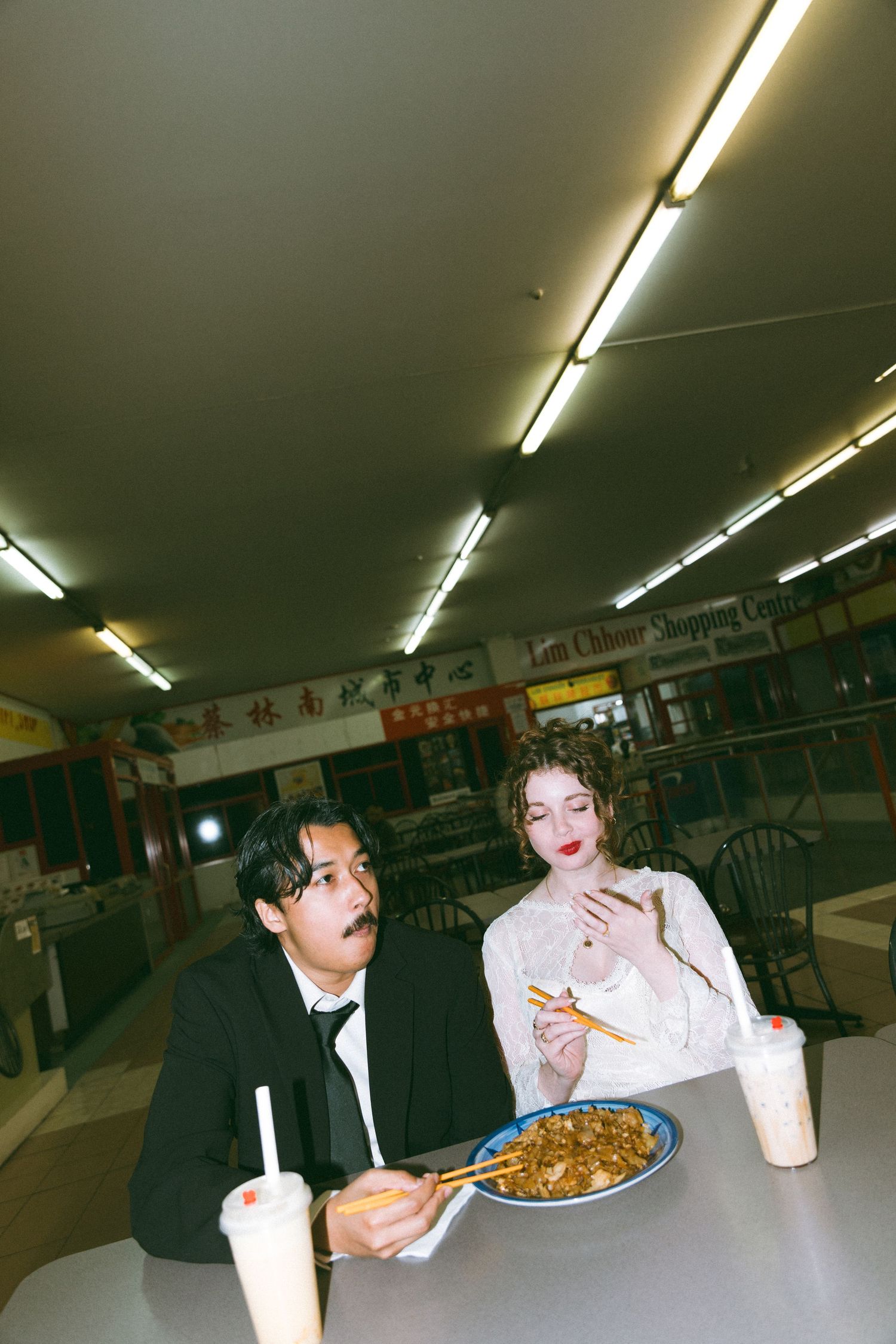 Bride and groom share noodles with bubble tea at a casual Asian shopping center restaurant.