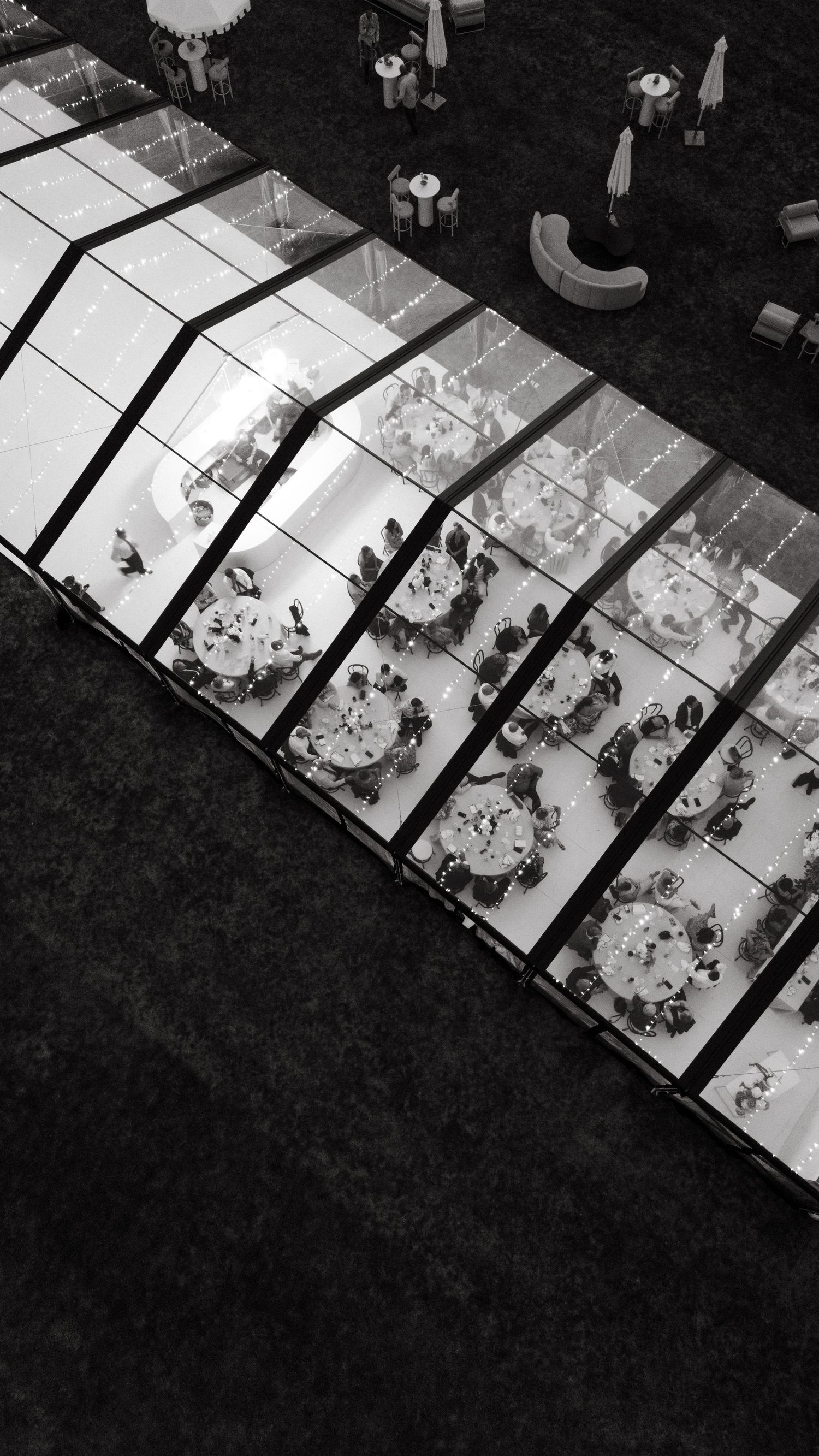 Aerial black and white view of guests seated at round tables inside a glass marquee wedding reception venue.