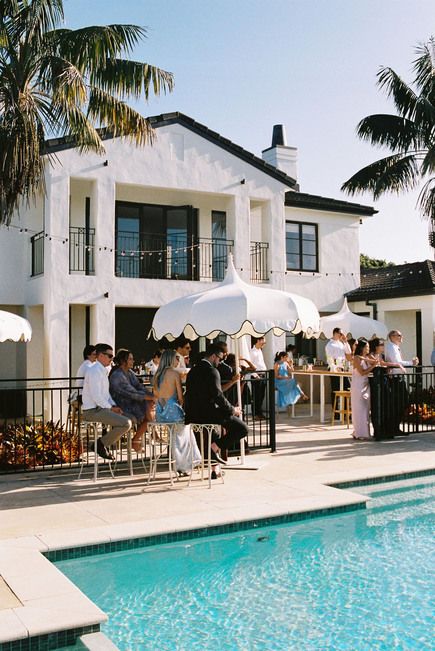 Wedding guests mingling poolside at elegant white villa with palm trees and striped umbrellas.