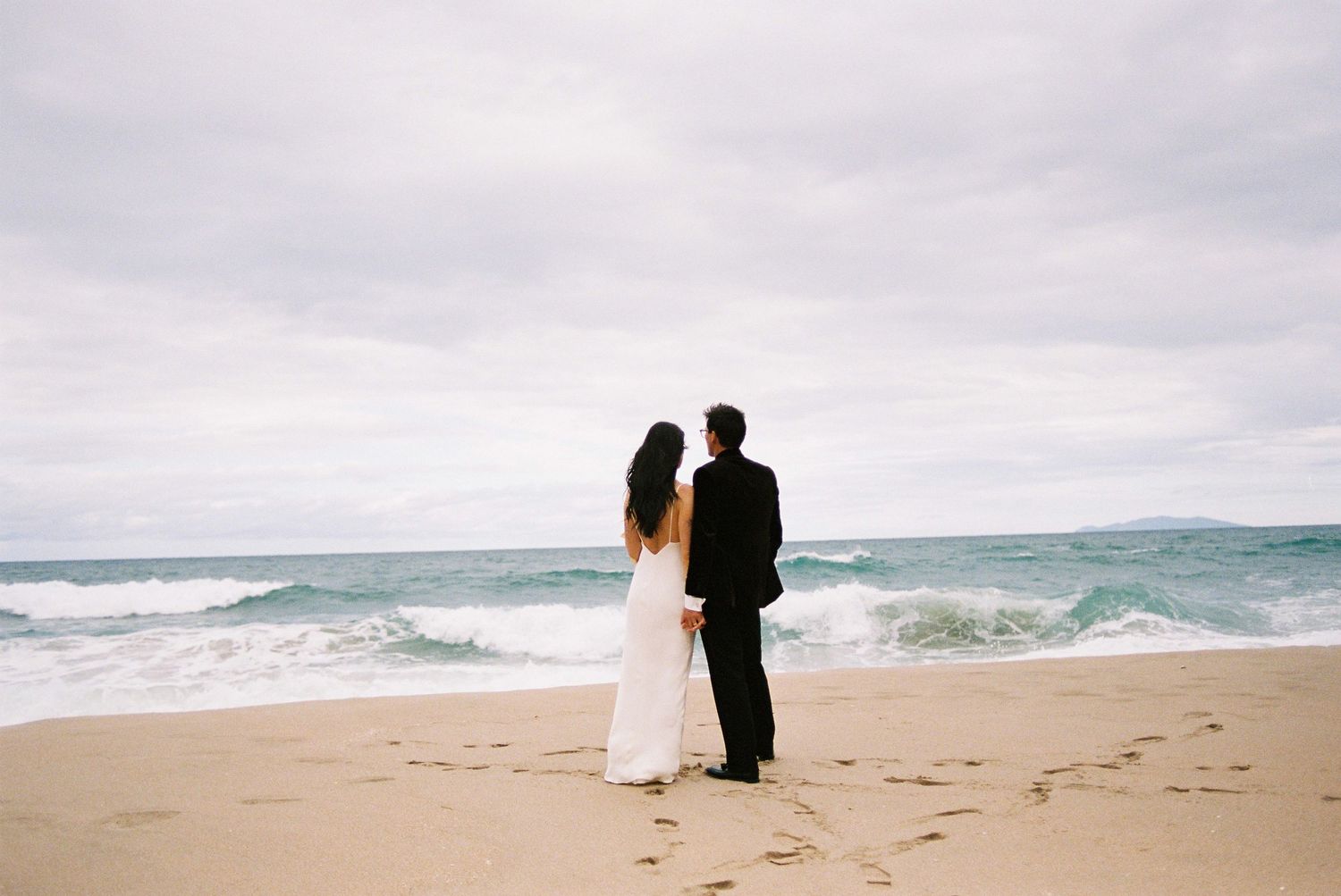 Bride and groom stand together on a sandy beach facing ocean waves under a cloudy sky.