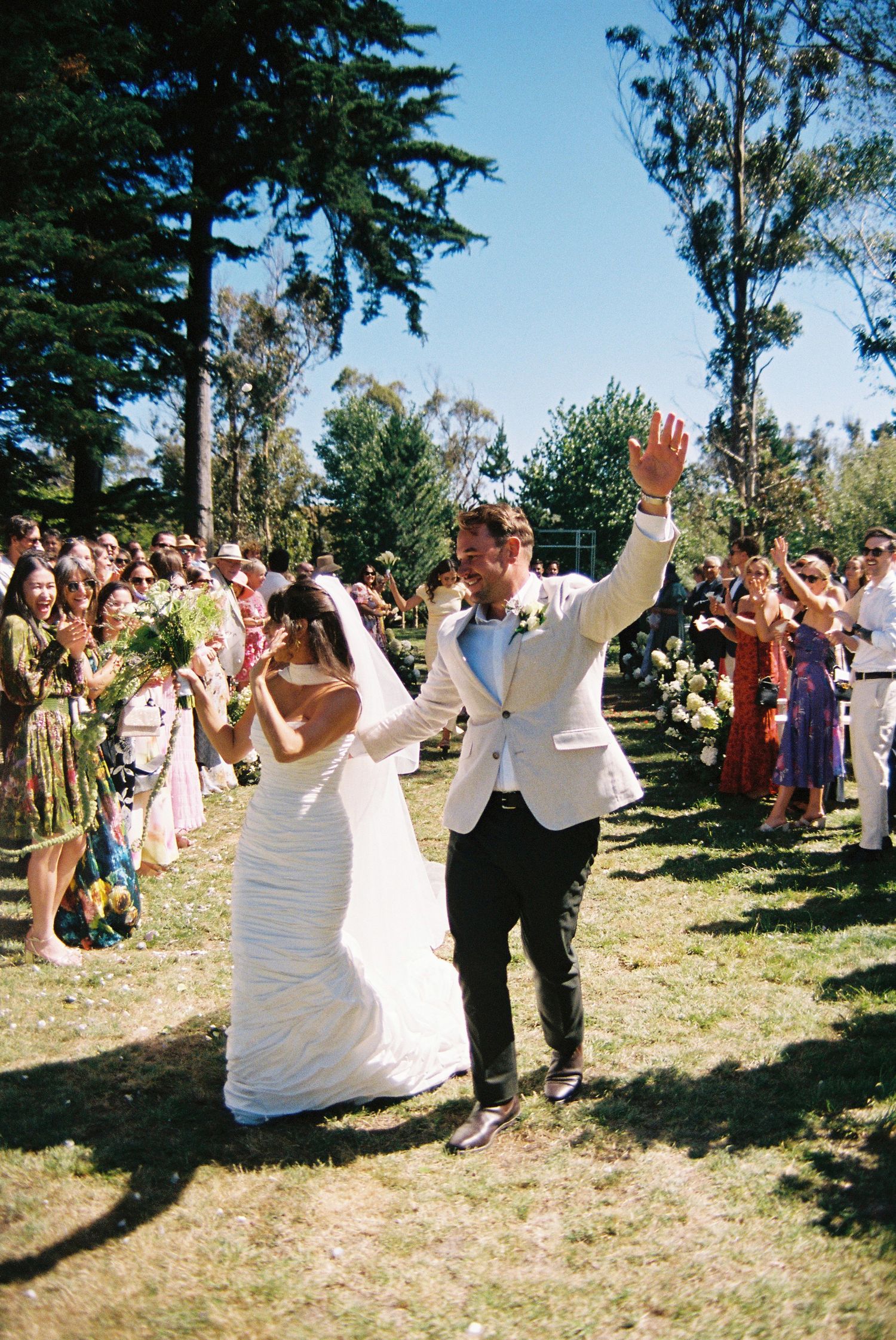 Joyful newlyweds walking back down the aisle outdoors, groom raising hand, guests cheering in sunny garden setting.