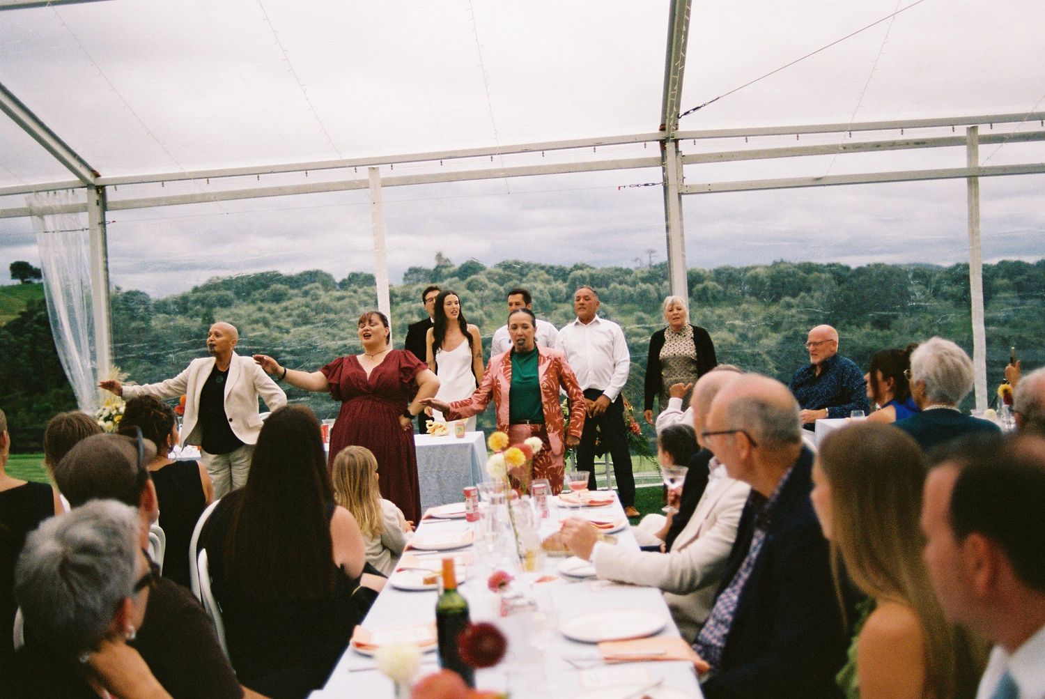 Wedding guests seated at long tables inside clear marquee tent while bridal party stands and celebrates.