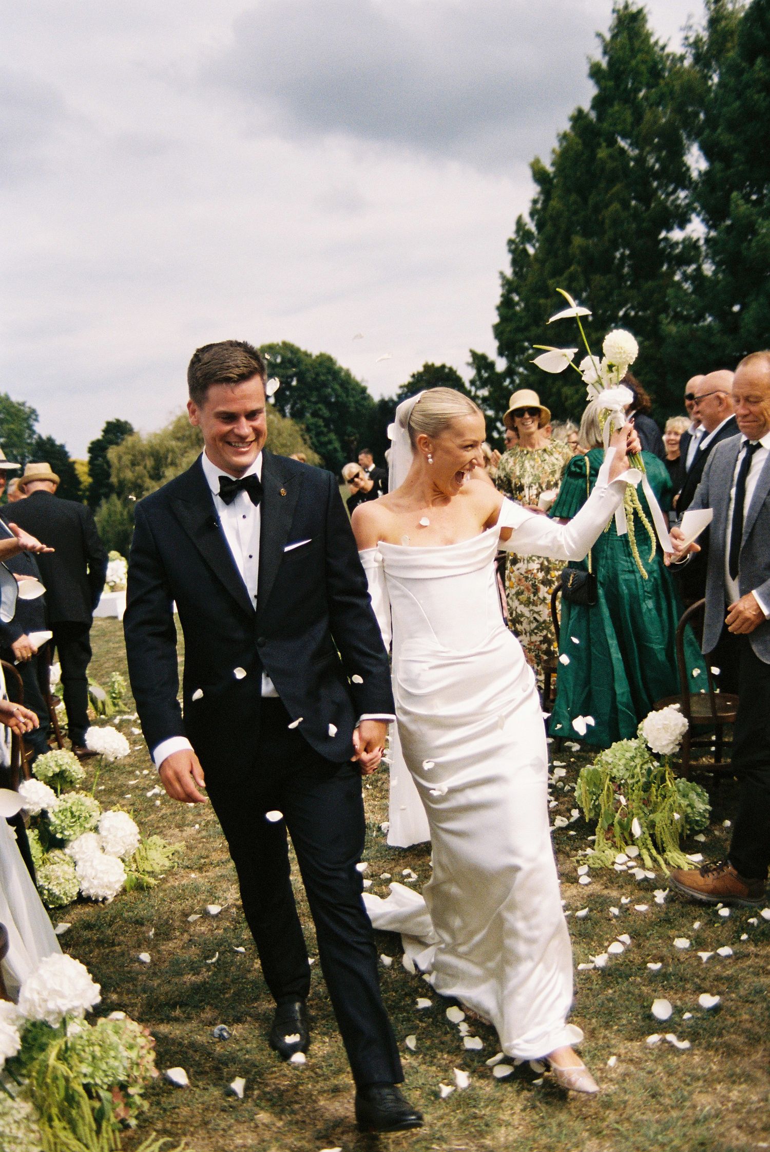 Bride and groom walking down the aisle after ceremony as guests throw white flower petals in outdoor wedding.