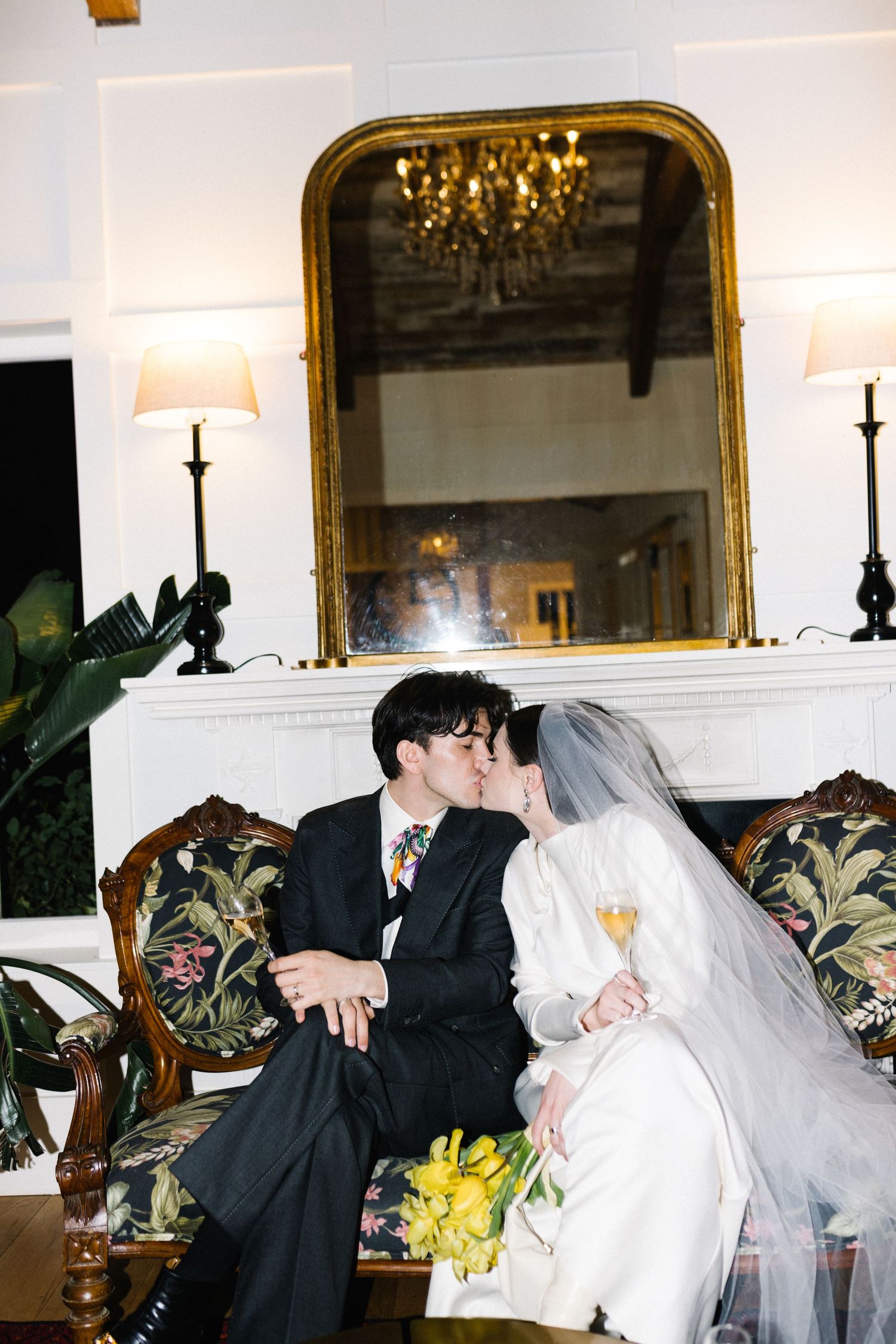 Bride and groom share a kiss on a floral sofa by a fireplace with a gold mirror, bride holding yellow flowers. new zealand photographer film