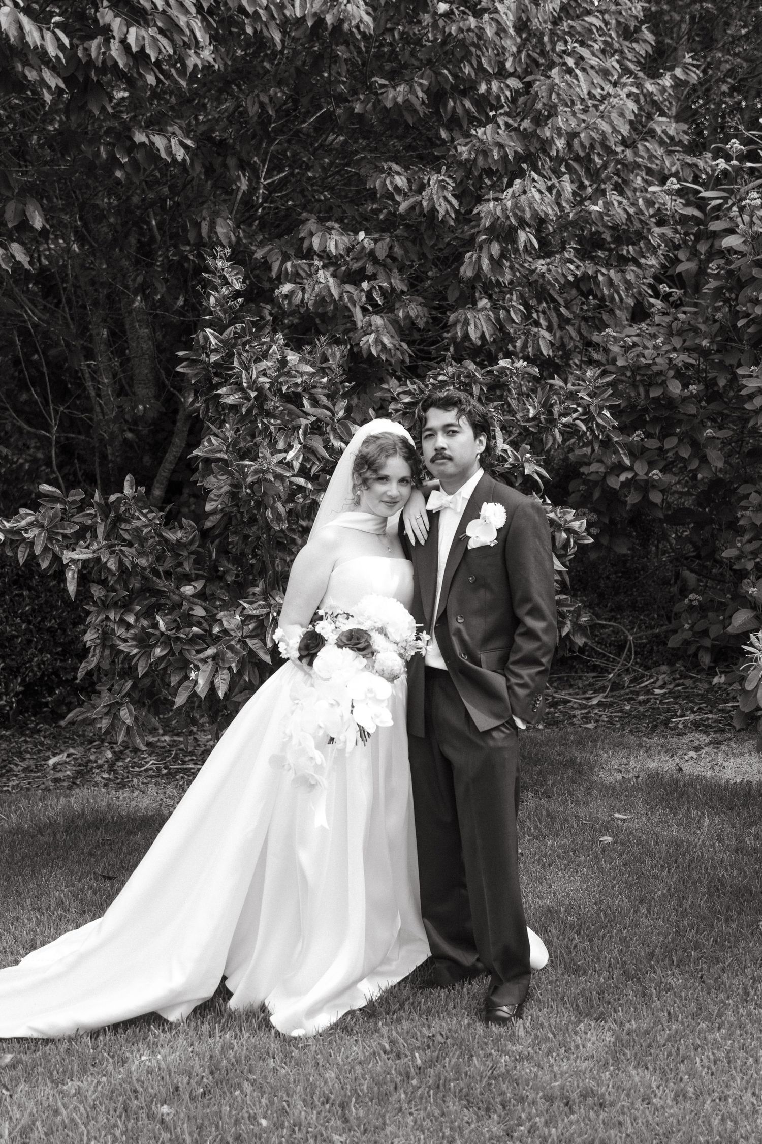 Black and white photo of bride in white gown and groom in tuxedo posing in garden.