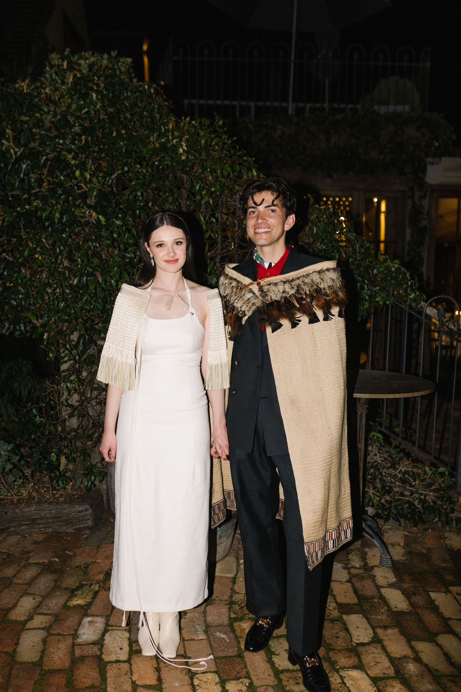 Couple posing outdoors at night, bride in white gown with fringe shawl, groom in black suit with decorative blanket wrap.