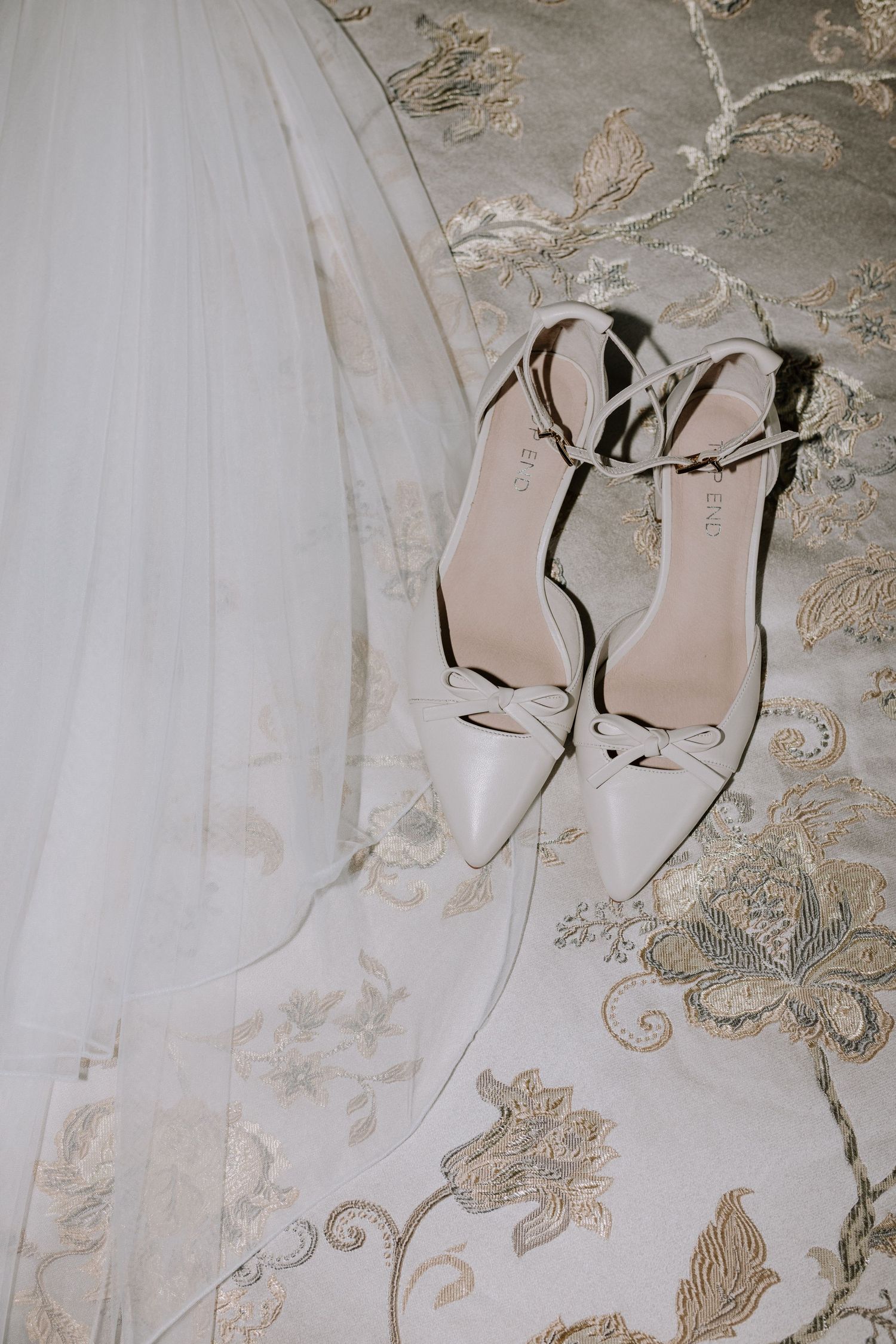 White bridal heels with bow detail and ankle straps rest on embroidered floral fabric.