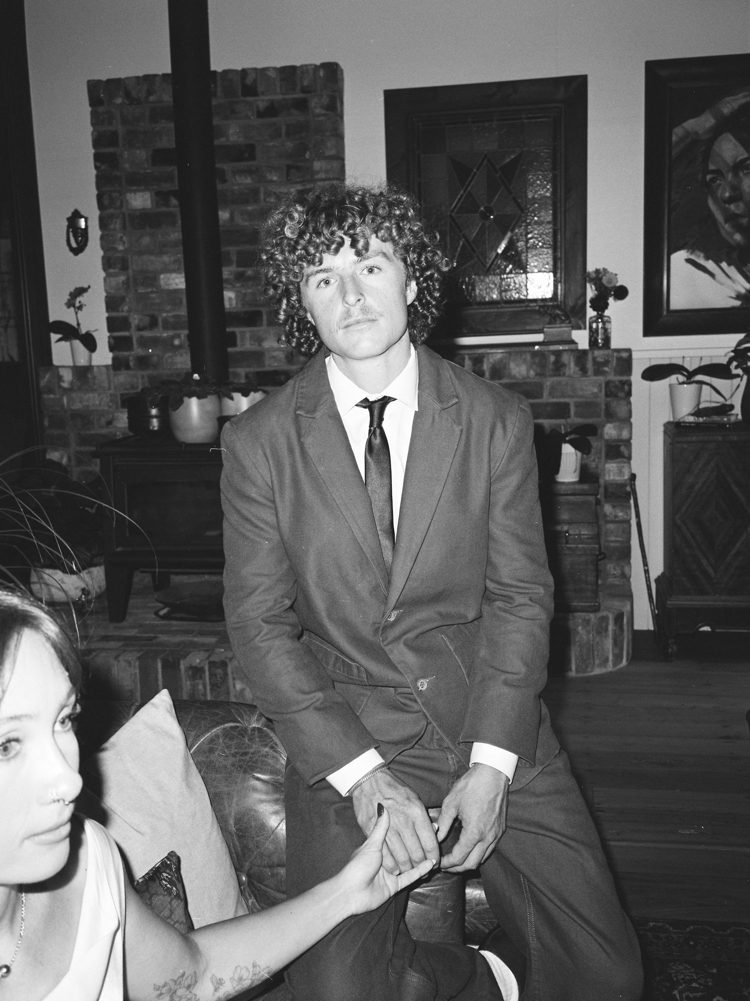 Young man with curly hair in a brown suit and tie sitting at an indoor wedding reception in black and white.