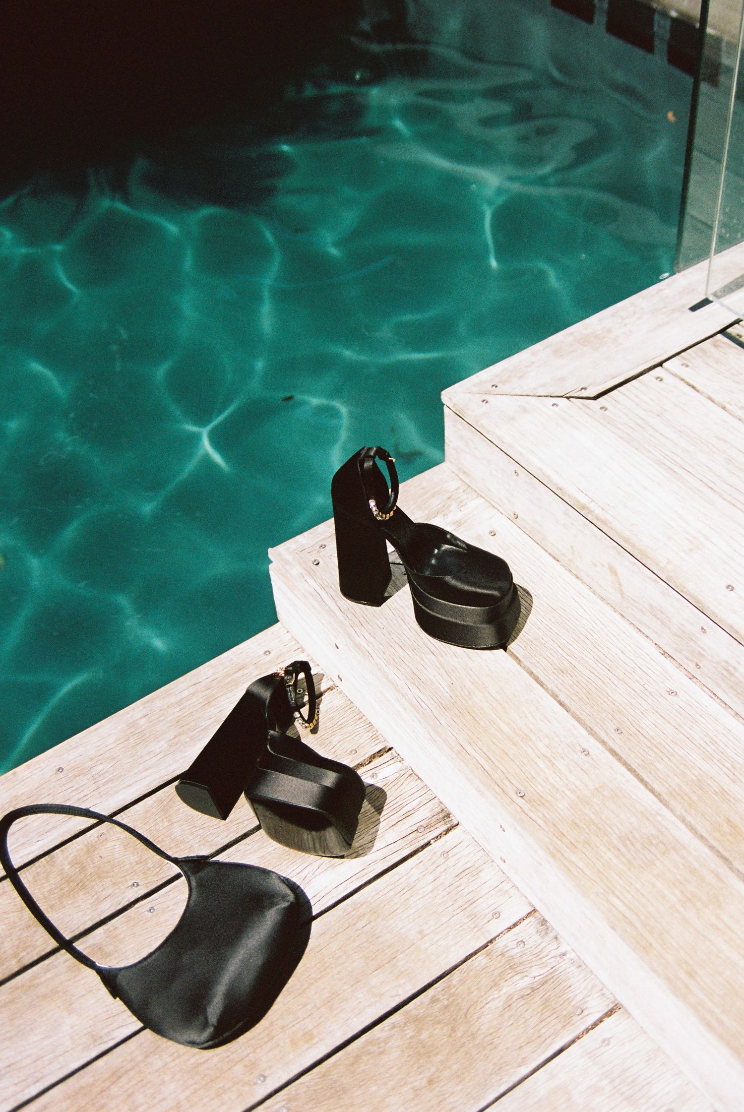 Black platform heels and small bag resting on wooden poolside deck beside turquoise water.