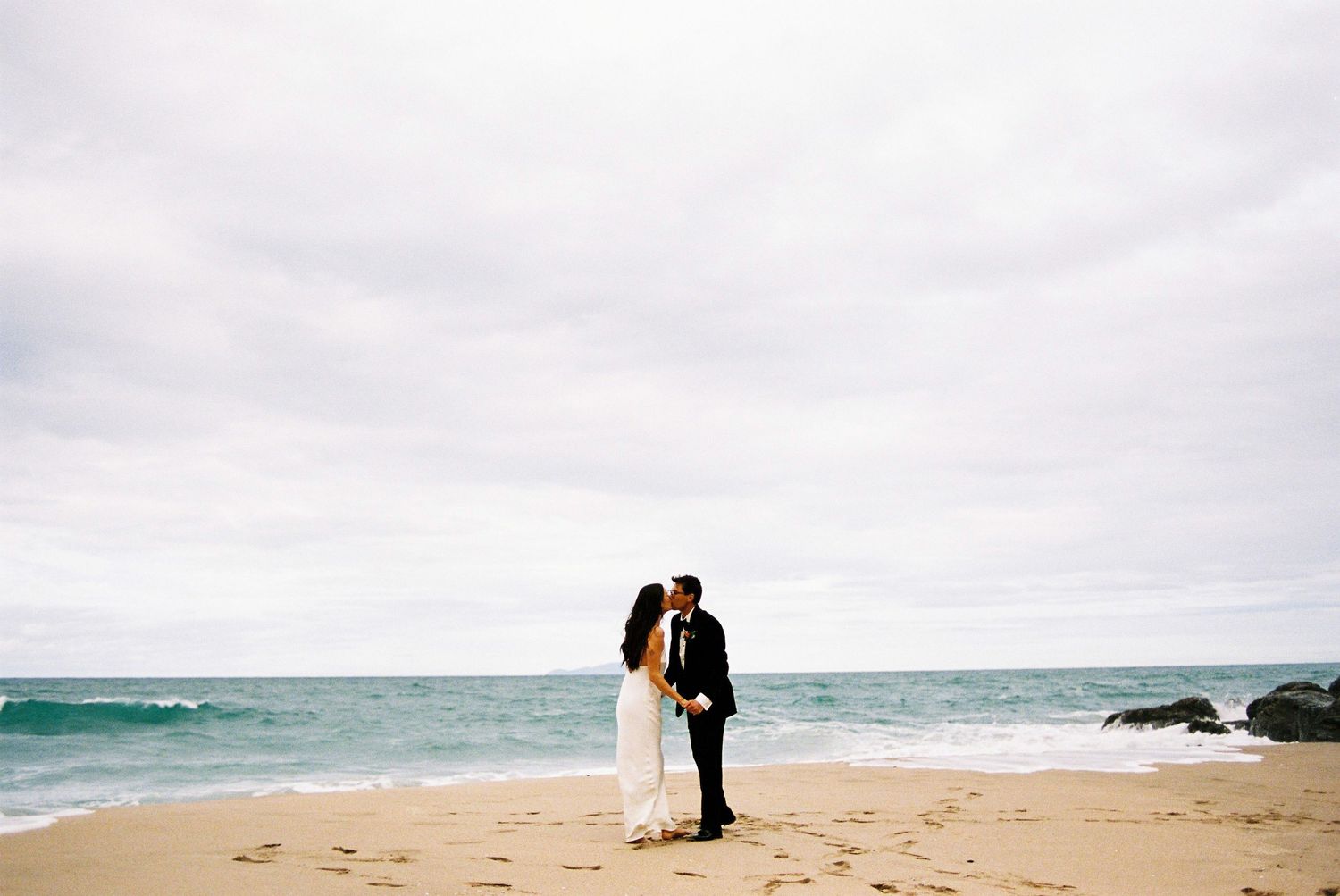 Bride and groom sharing a kiss on a sandy beach with ocean waves and cloudy sky behind them.