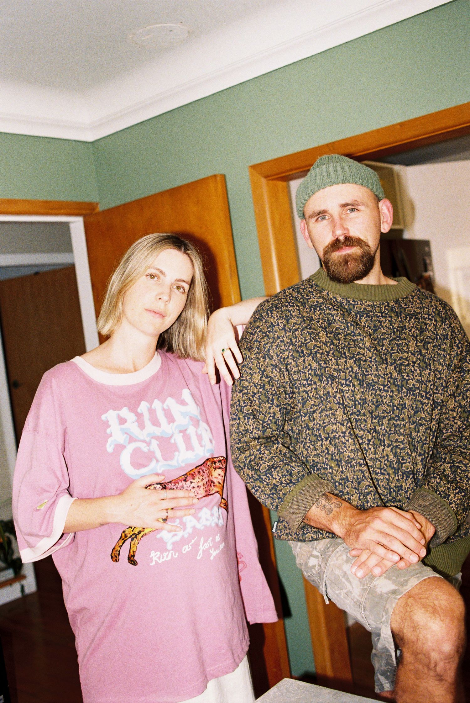 Couple posing casually indoors, woman in pink Run Club tee, man in patterned sweater and beanie.