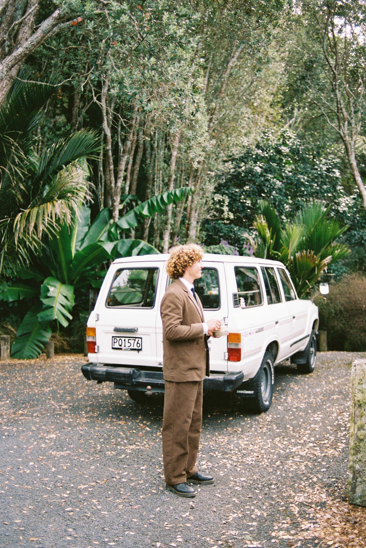Curly-haired groom in brown suit standing beside a vintage white Toyota Land Cruiser on a leafy driveway.