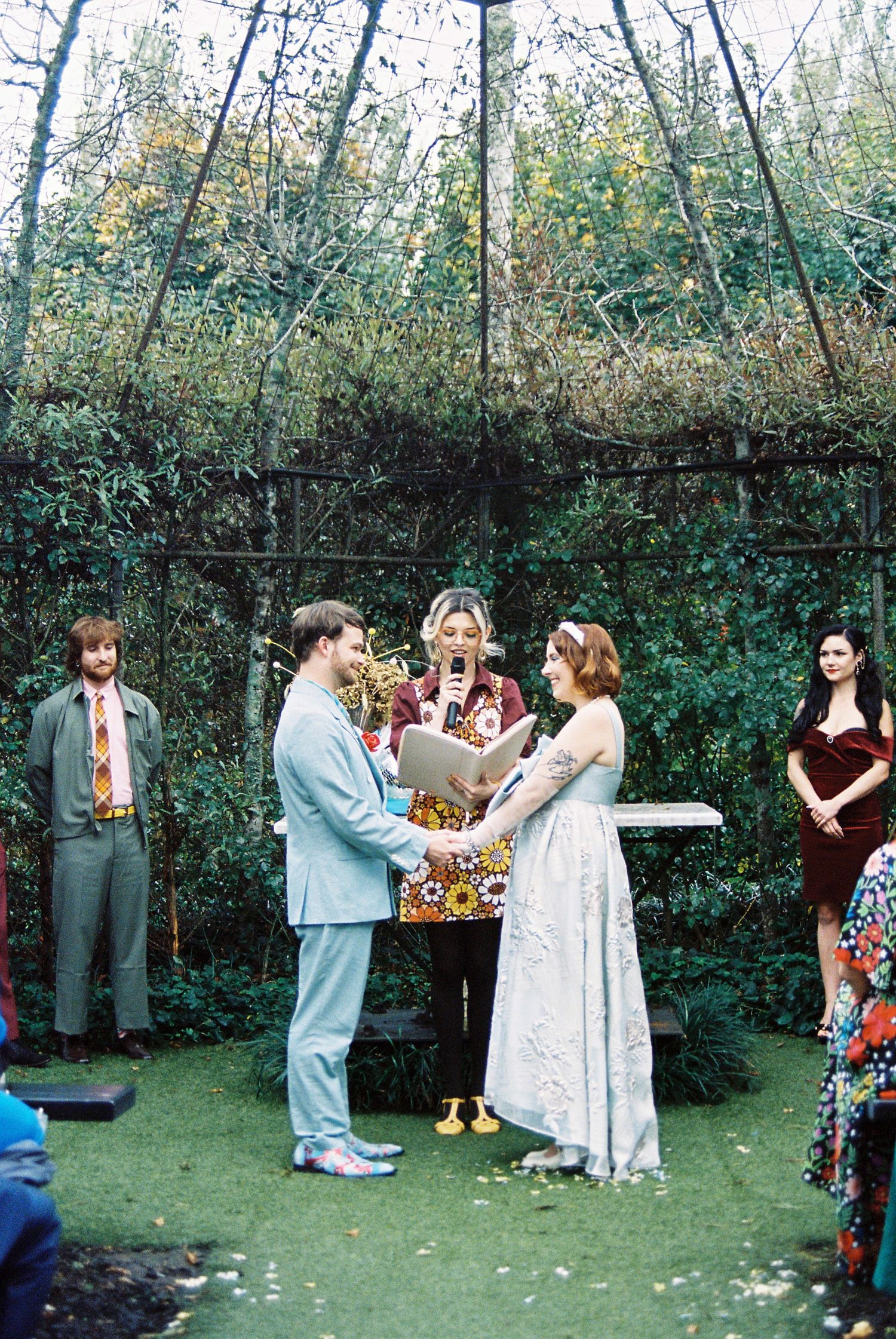 Couple exchanges vows at outdoor wedding ceremony surrounded by lush greenery with officiant.