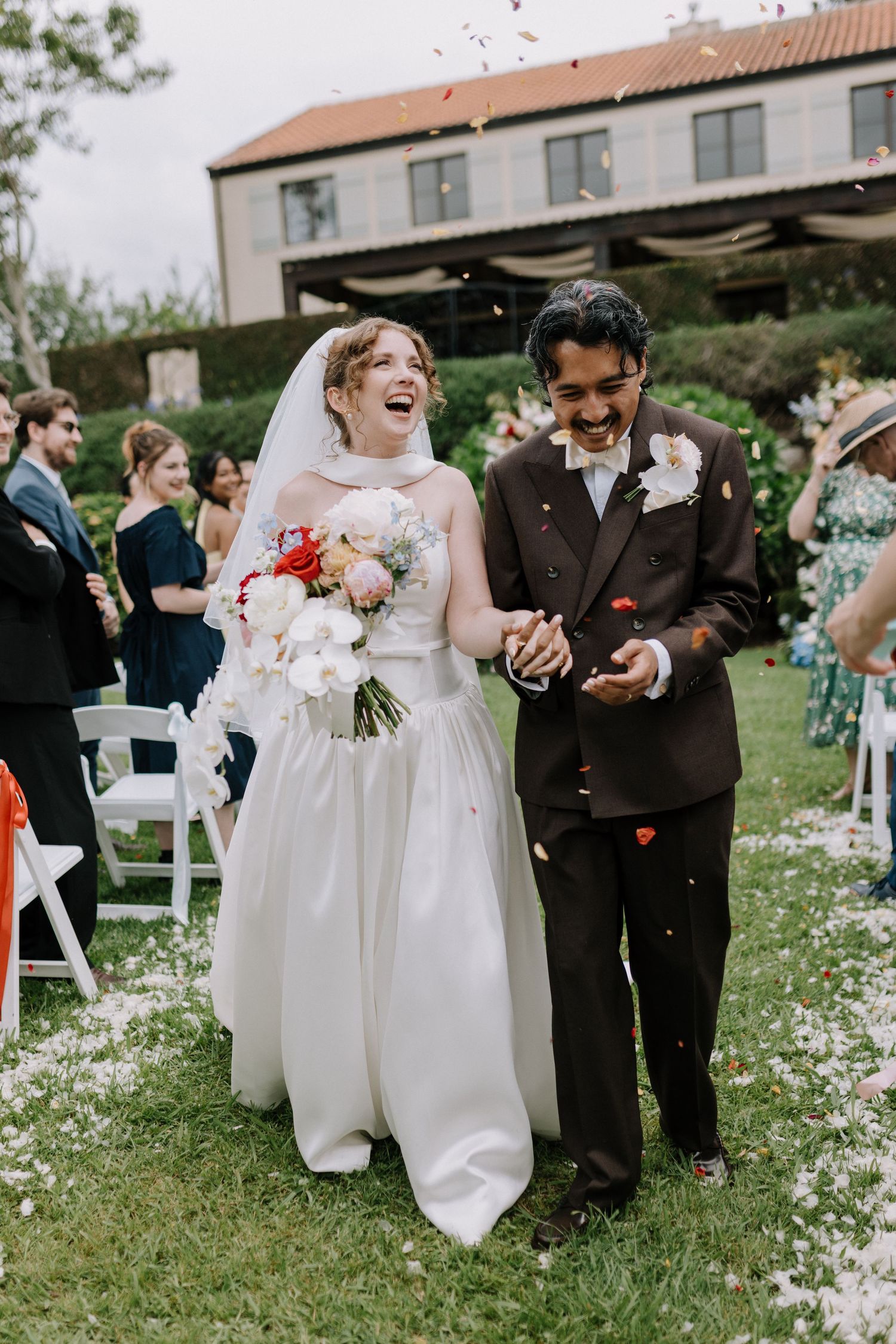 Laughing newlyweds walk down aisle through confetti shower at outdoor garden wedding ceremony.