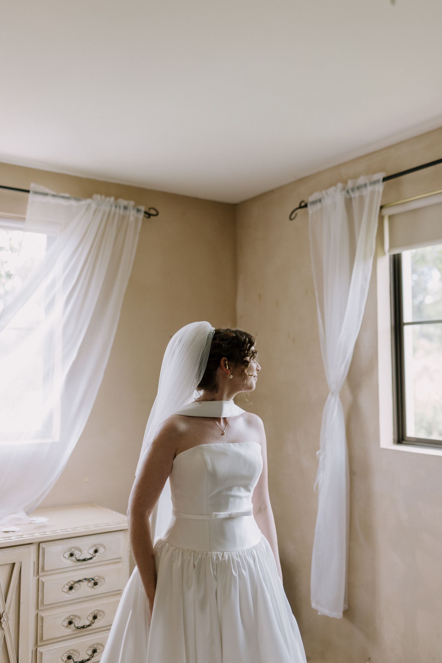 Bride in strapless white gown and veil stands by window in soft natural light.