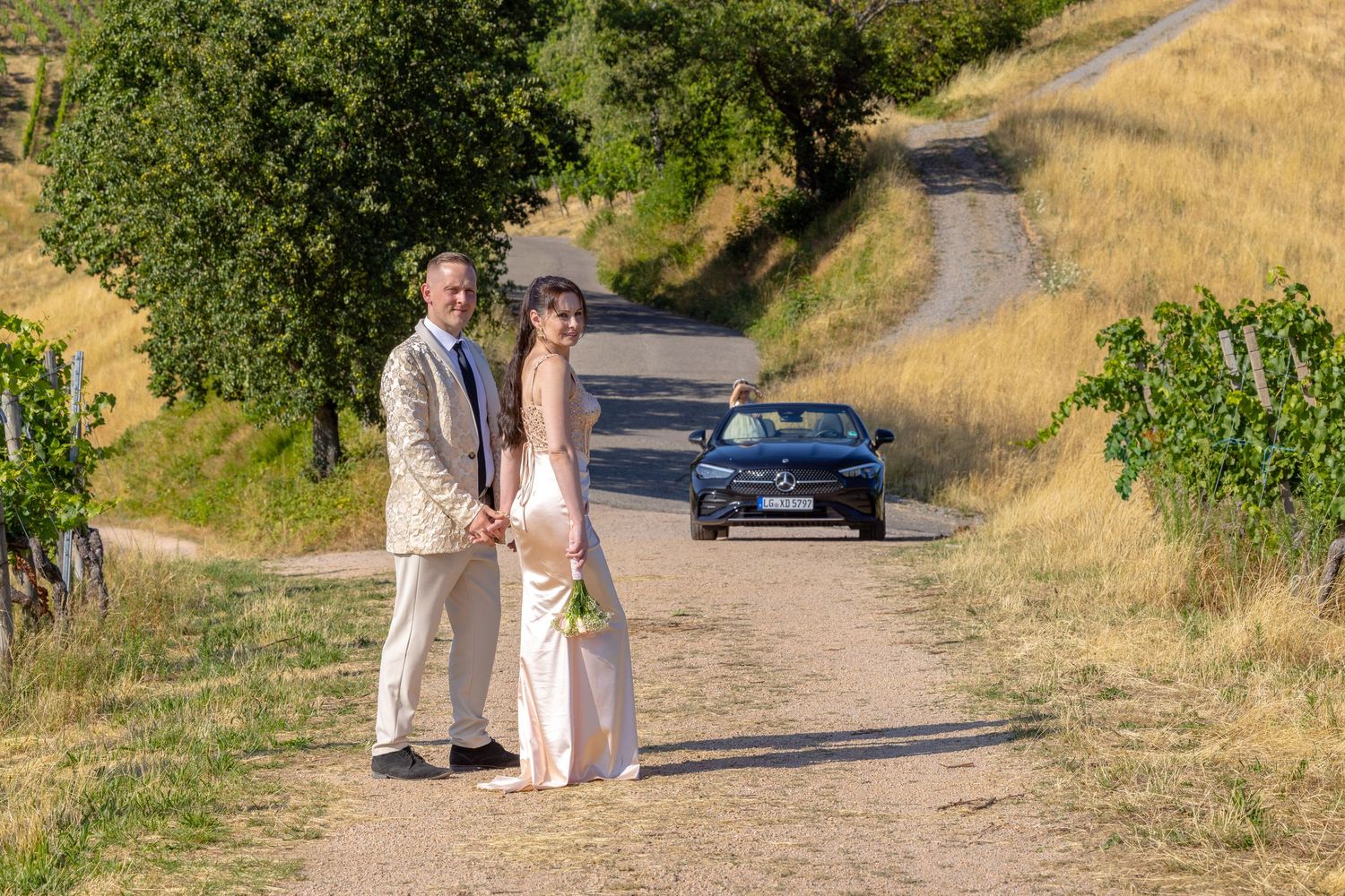 Brautpaar steht auf einem Weg vor Weinbergen und geparktem Cabrio