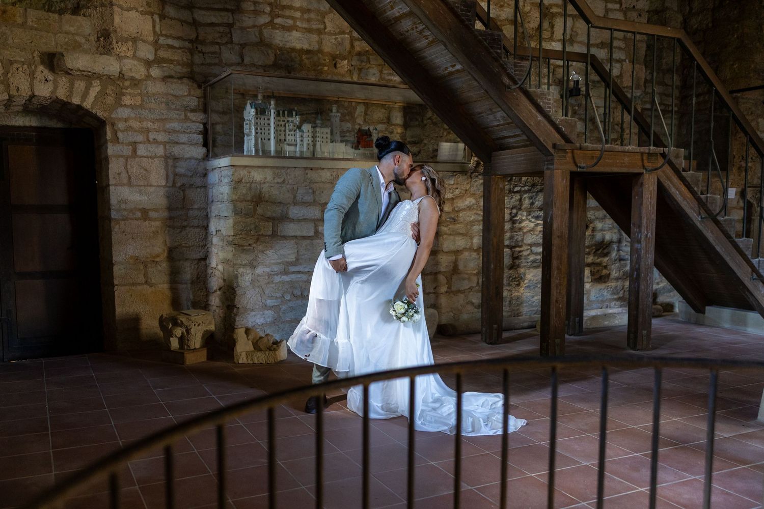 Naomi und Sergiu küssen sich unter einer alten Holztreppe im historischen Innenraum.