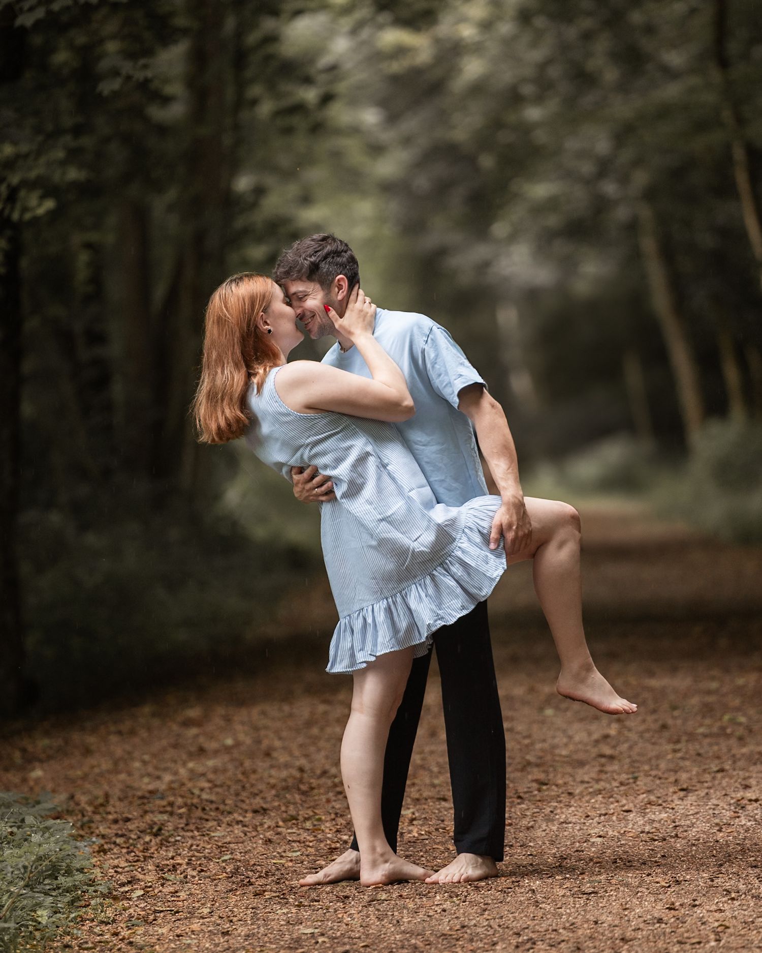 A couple shares a romantic kiss on a forest path, the man lifting the woman in a light blue dress.