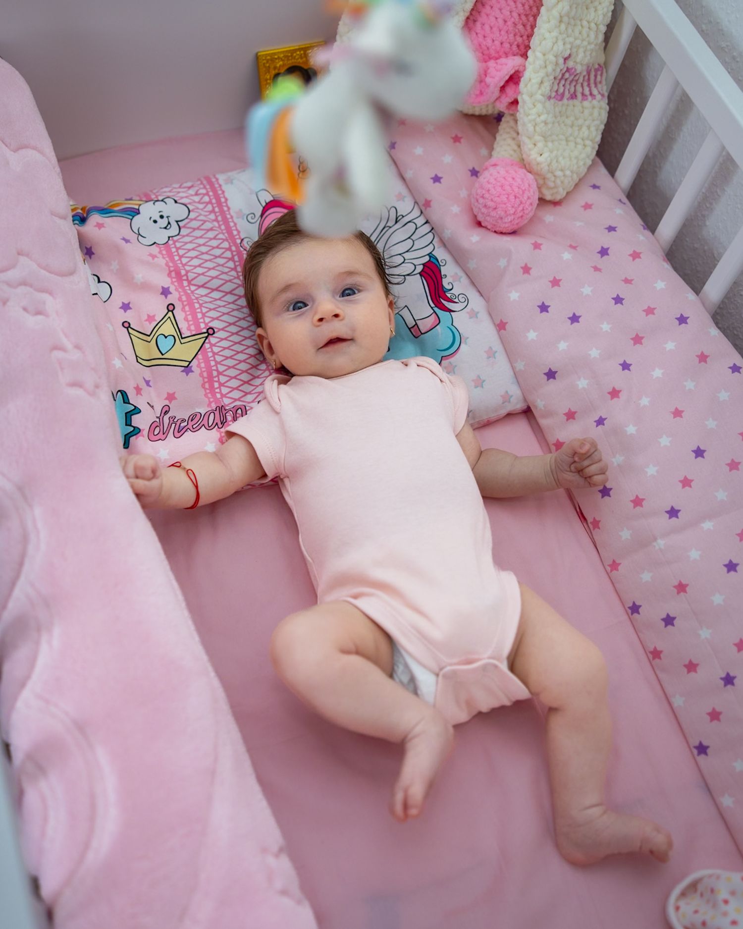 A cute baby girl in a pink onesie lying in a pink crib surrounded by colorful toys and star-patterned bedding.