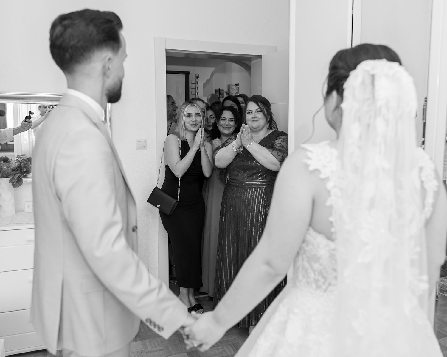 Black and white photo of bride and groom holding hands as bridesmaids react with joy in doorway at wedding.