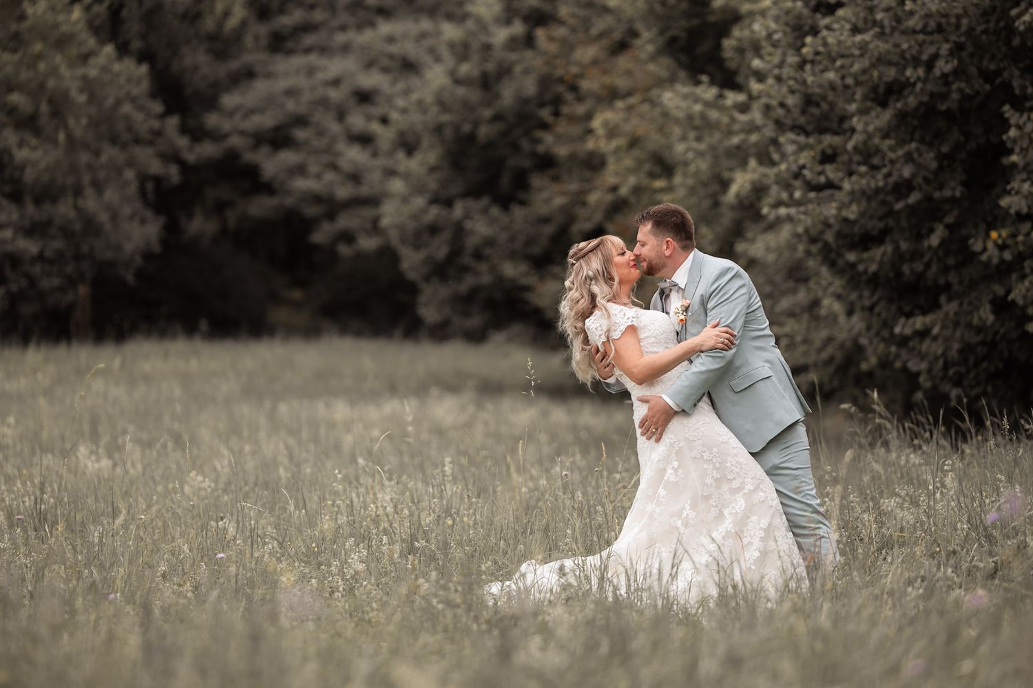 Bride and groom sharing a romantic kiss in a lush green meadow, with the groom dipping the bride in her lace wedding gown.