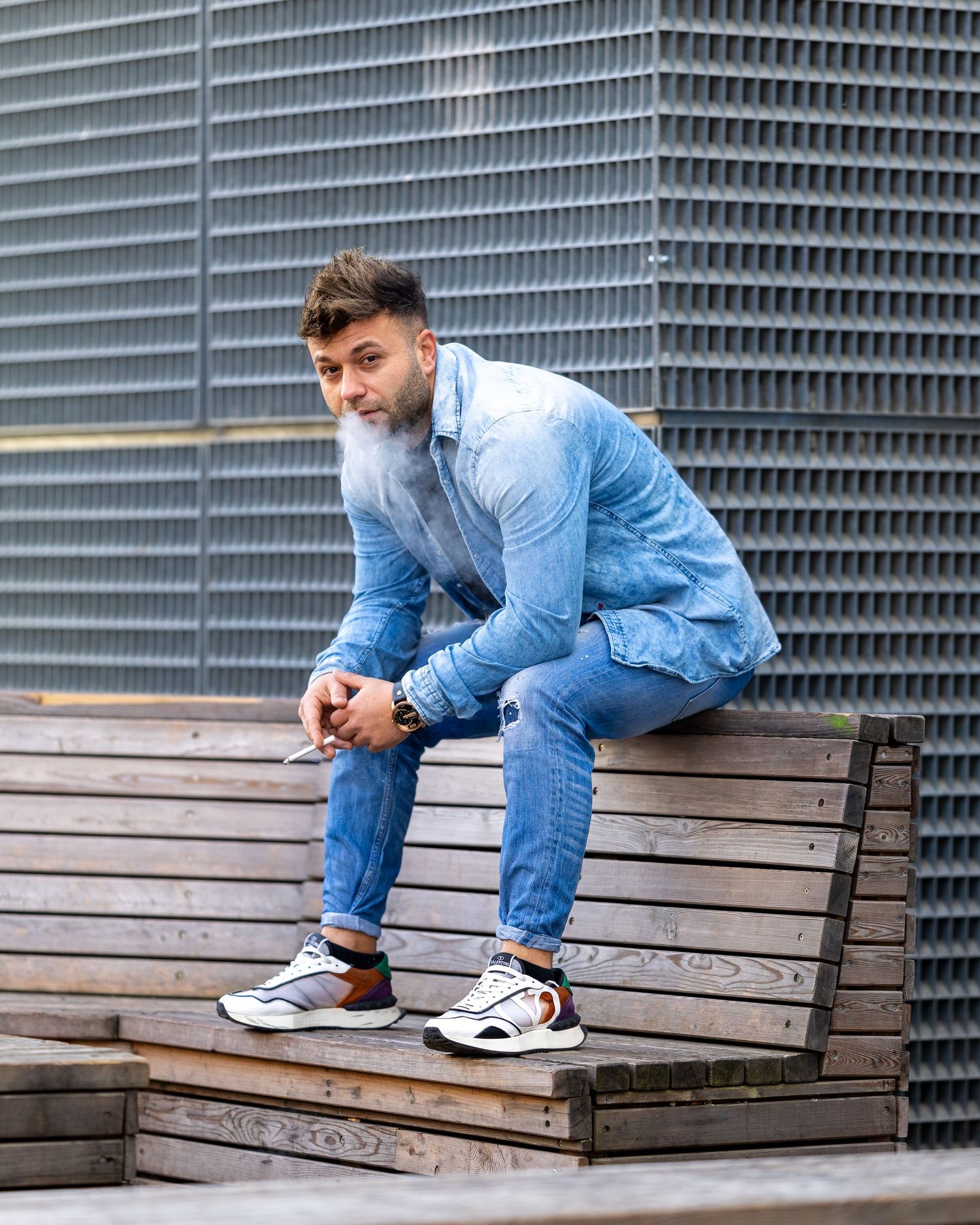 Man in blue denim outfit sitting on wooden steps outdoors, wearing white sneakers, with a stylish beard.
