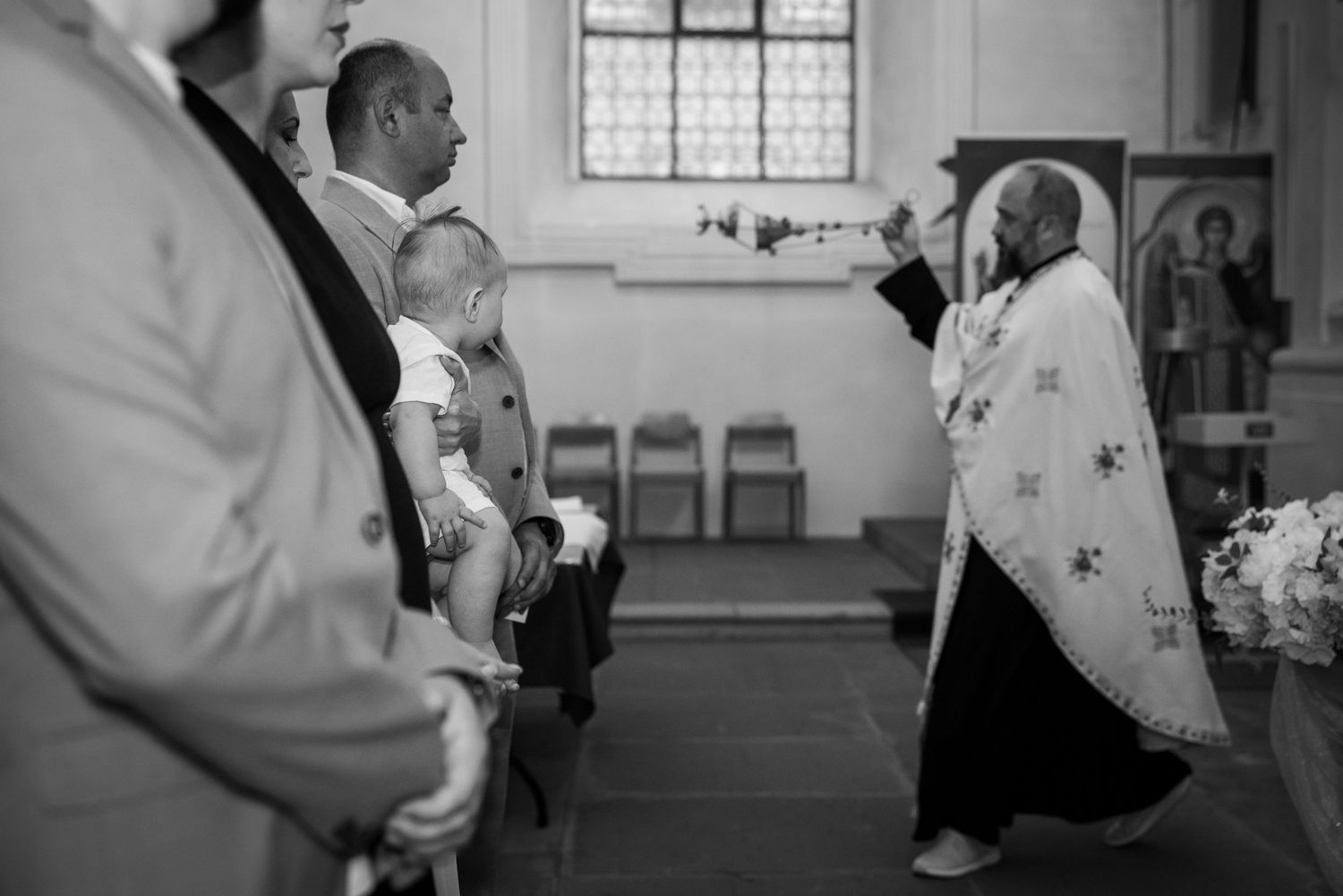 Aris auf dem Arm eines Mannes, während der Priester das Weihrauchfass schwenkt; Schwarz-Weiß-Aufnahme.