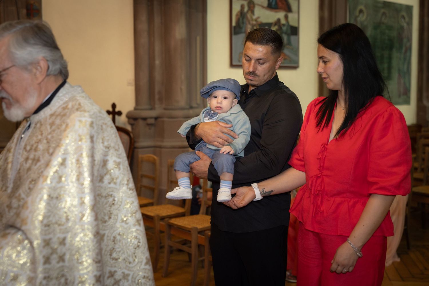 Carlo wird von einem Mann neben einer Frau in Rot gehalten, während der Priester die Taufe in Straßburg begleitet.