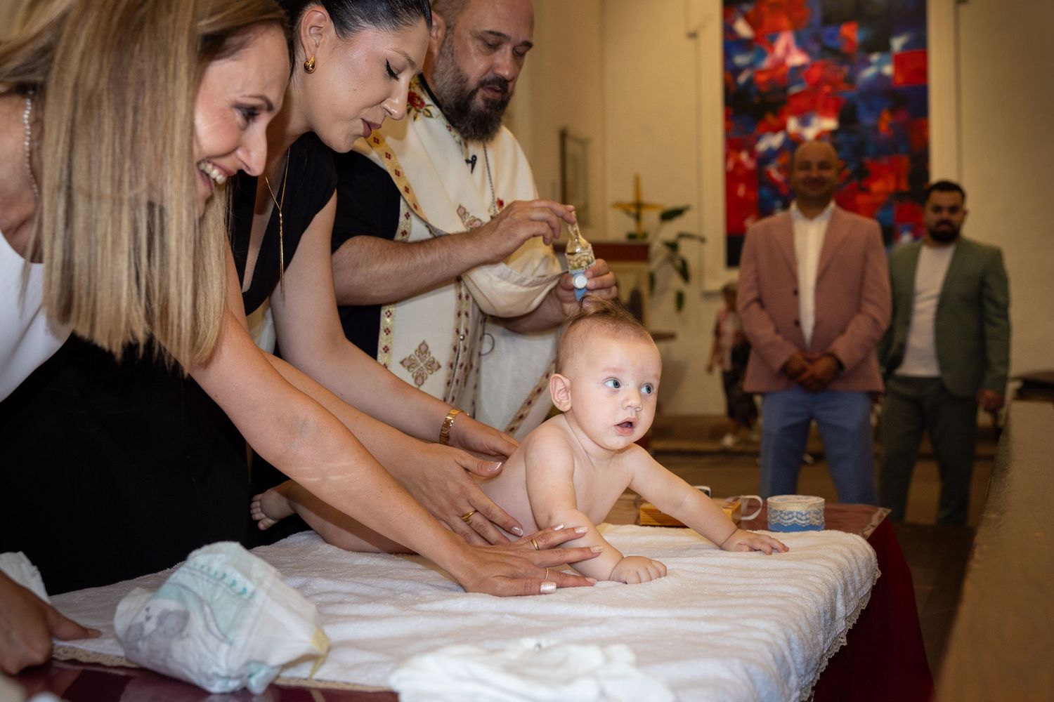 Zwei Frauen stützen Aris auf einer weißen Unterlage, während der Priester die Salbung vorbereitet.