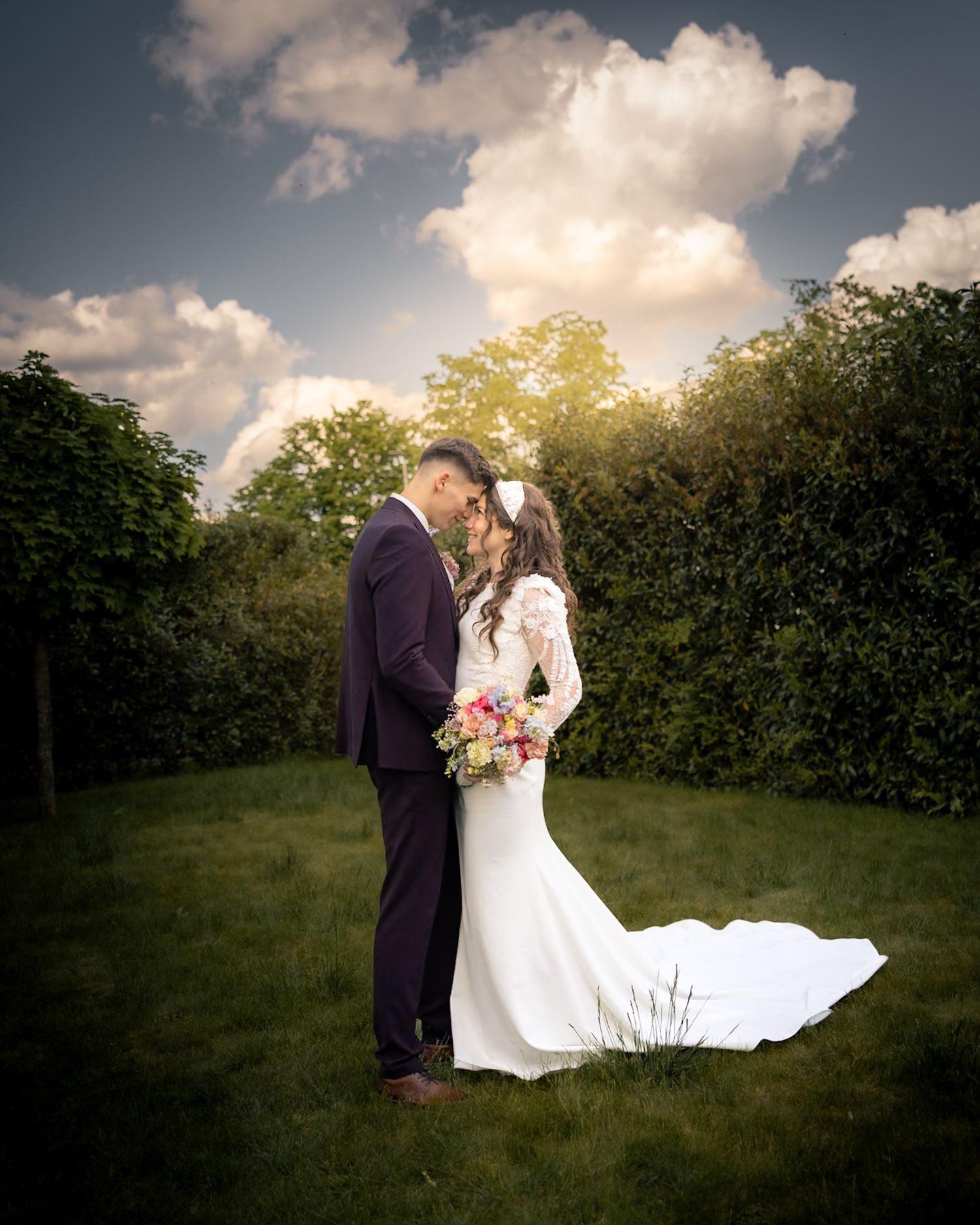 Bride and groom embrace in lush garden with dramatic cloudy sky backdrop.