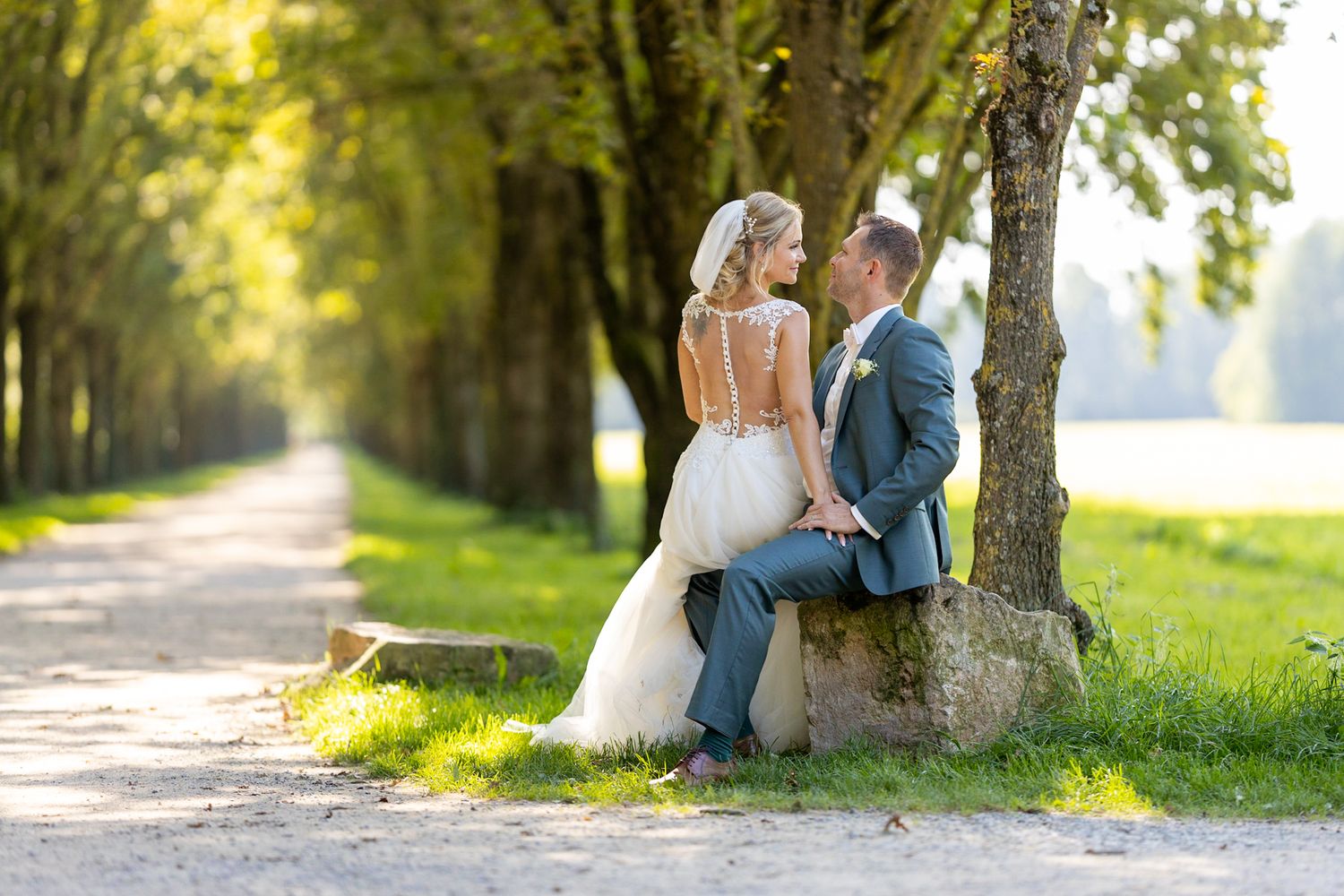 Bride and groom sitting together by a tree on a scenic tree-lined path on their wedding day.