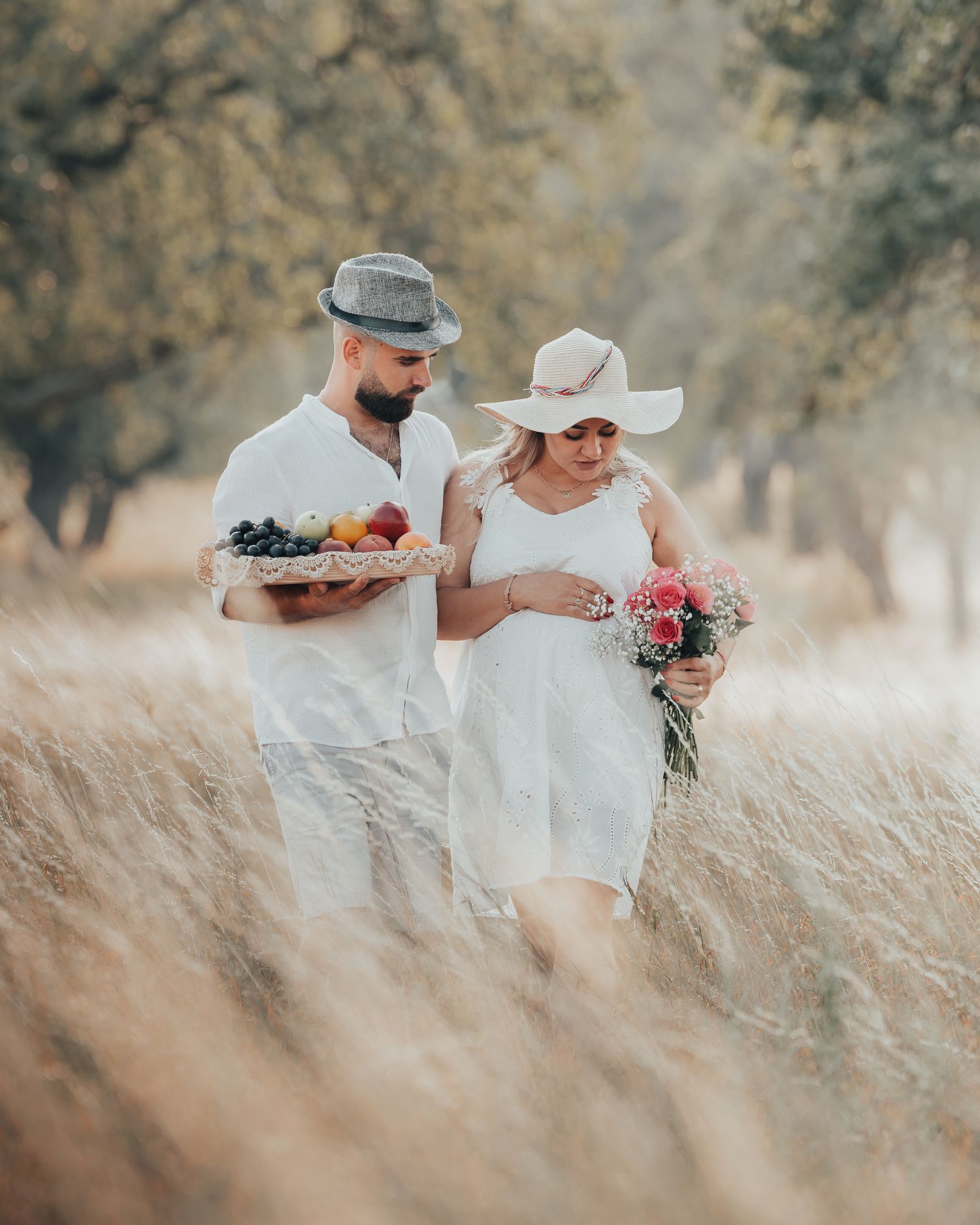 Couple in white outfits walking through golden fields, man holding fruit basket, woman carrying pink flowers, wearing hats.