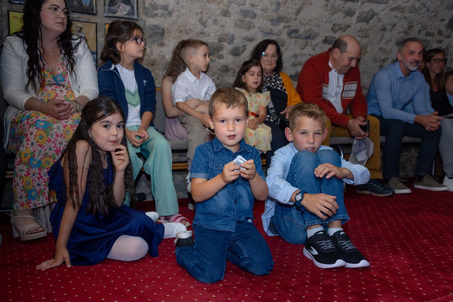 Kinder sitzen auf dem roten Teppich und verfolgen die Taufzeremonie, während Erwachsene hinter ihnen stehen
